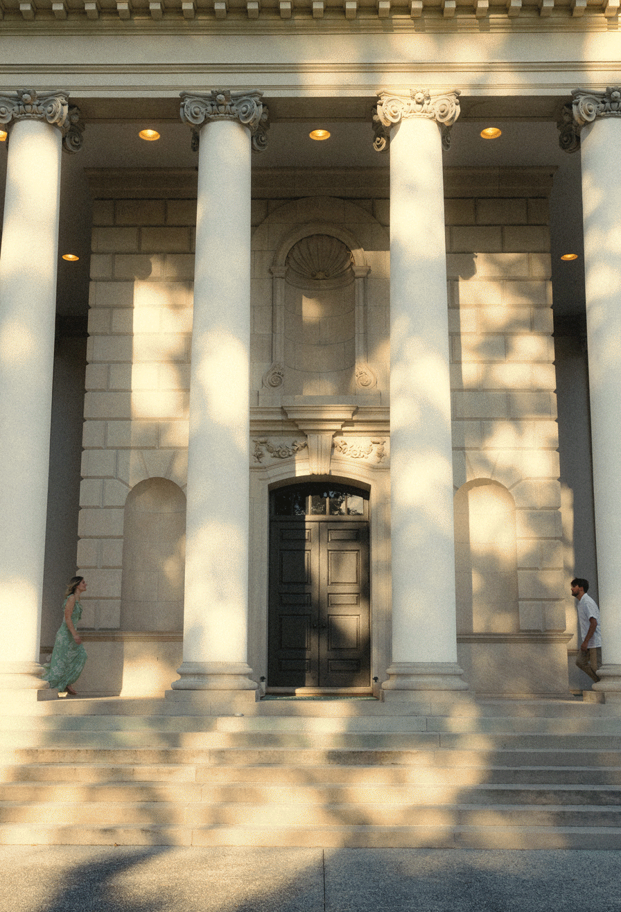 Classical building entrance with tall Corinthian columns casting dappled shadows on white stone facade and steps.