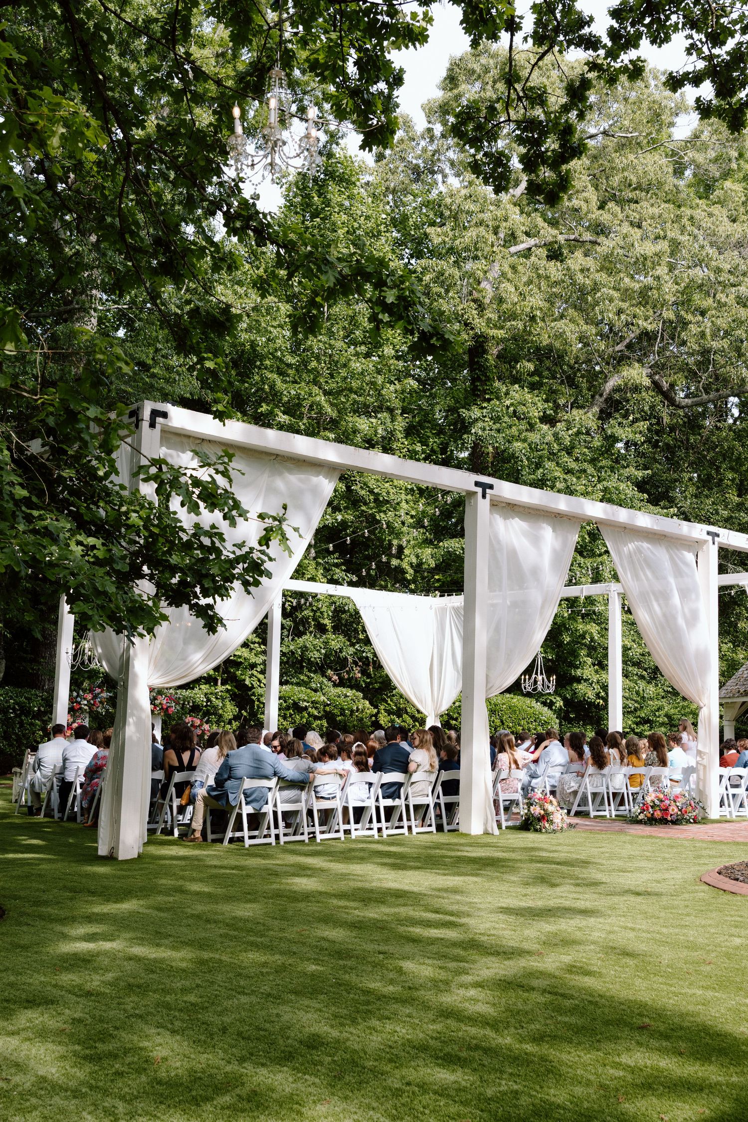 Outdoor wedding ceremony under white draped pergola surrounded by lush green trees with guests seated on white chairs.