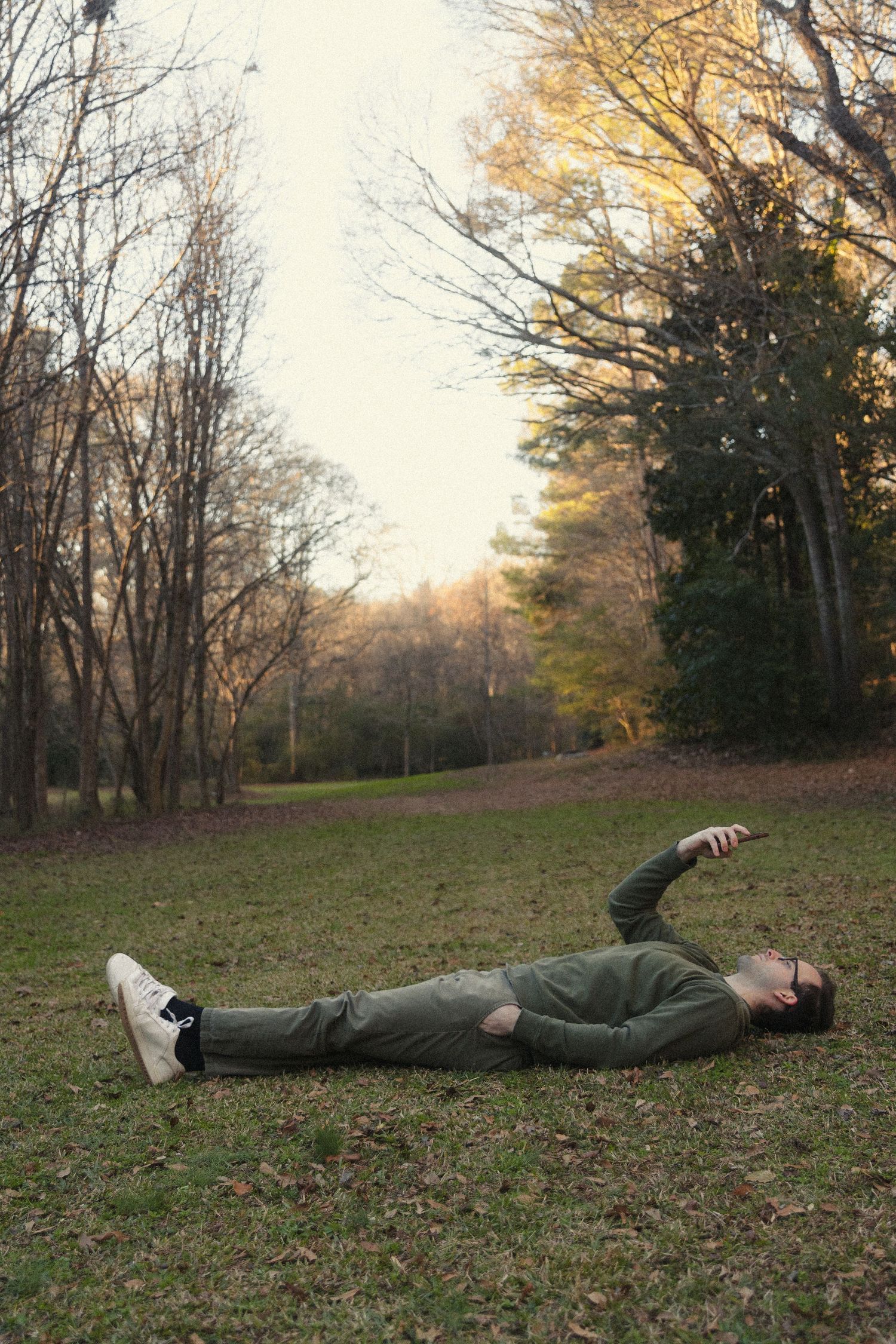Man lying on grass in autumn park wearing green sweater and white sneakers, surrounded by colorful fall trees.