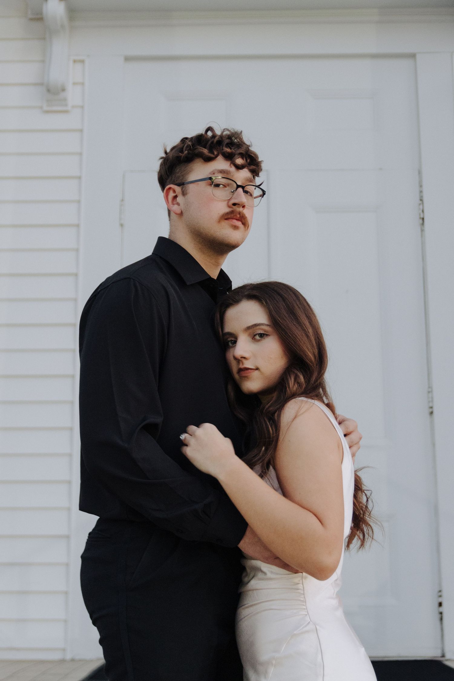 A romantic couple embraces against a white door, with them dressed in contrasting black and white outfits.
