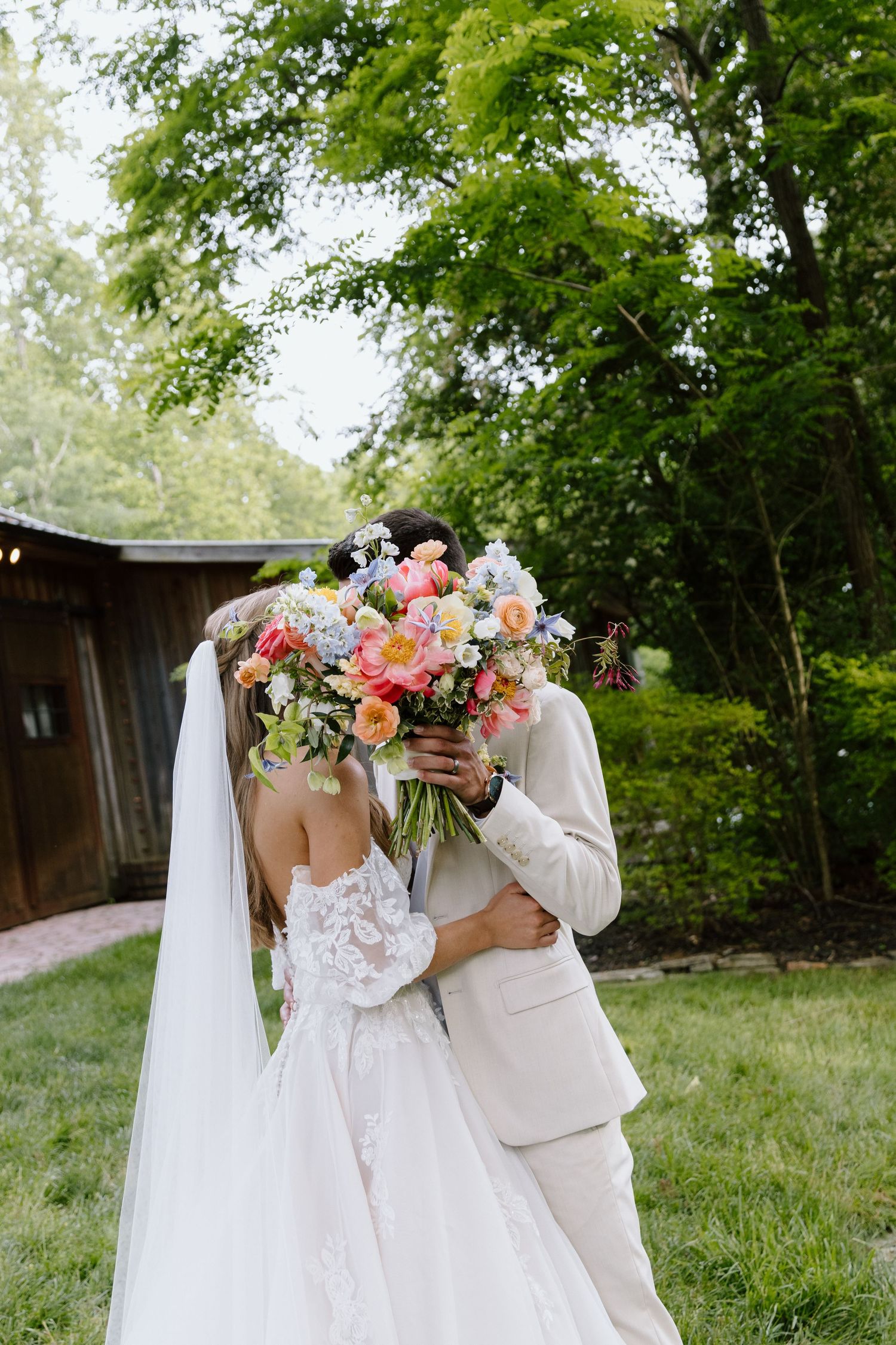 Bride and groom kissing behind colorful wedding bouquet in lush garden setting.