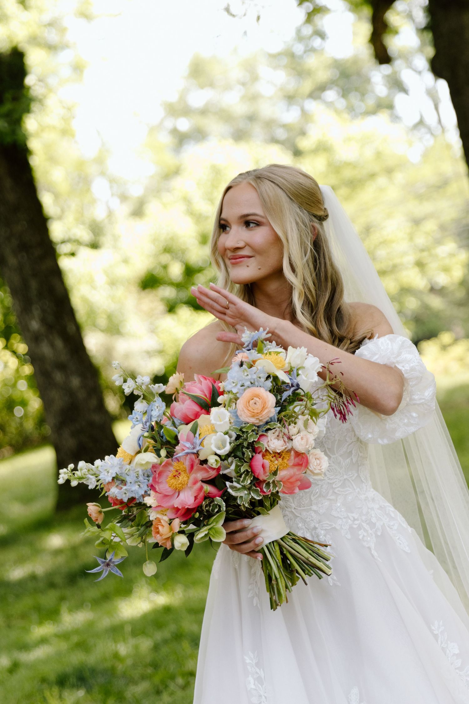 Smiling bride in white off-shoulder gown holds colorful spring bouquet outdoors in sunlit garden.