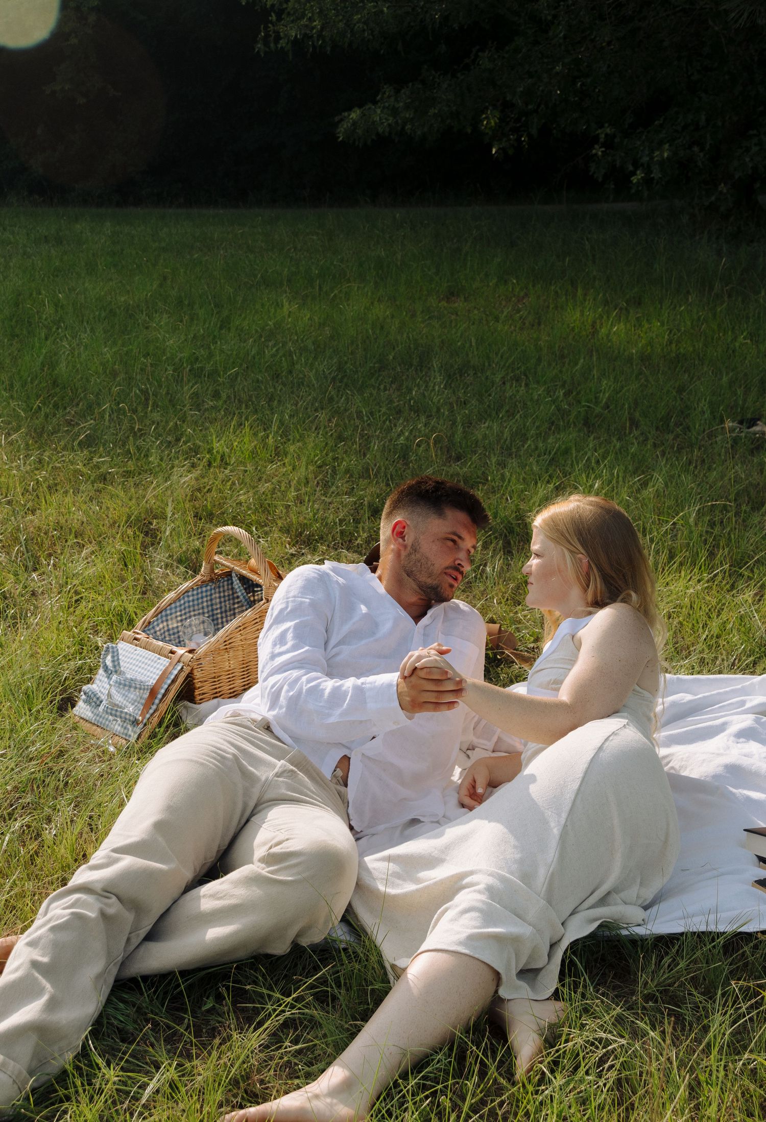 A couple enjoying a romantic summer picnic on a white blanket in a grassy field with a wicker basket nearby.