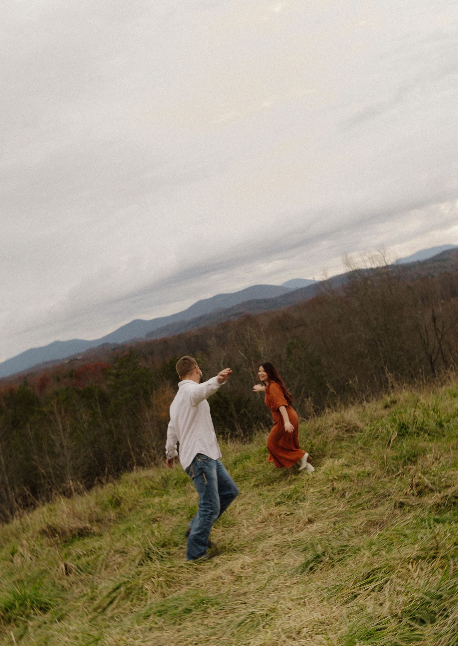 Two people walk along a grassy hillside path against a backdrop of misty mountains on an overcast day.