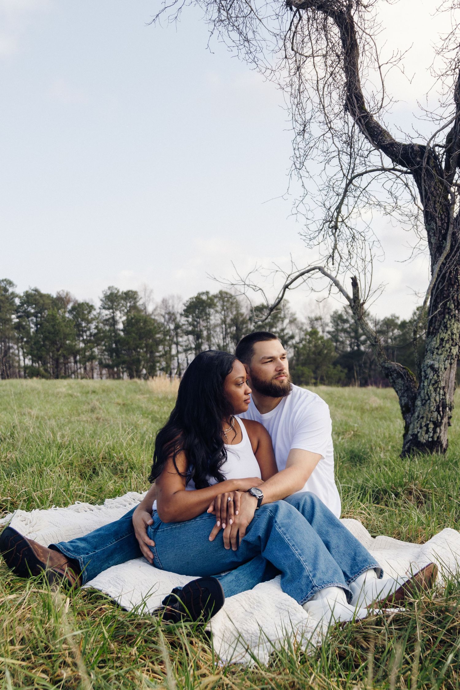 A couple sits intimately on a blanket in a grassy field under a bare tree on an overcast day.