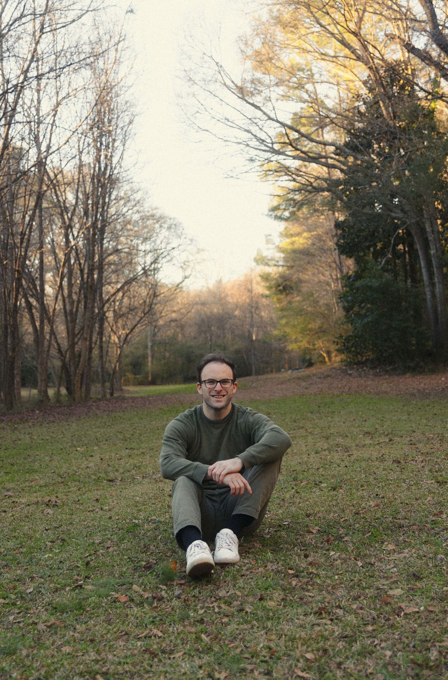 A person sits casually on grass in an autumn forest clearing during golden hour.