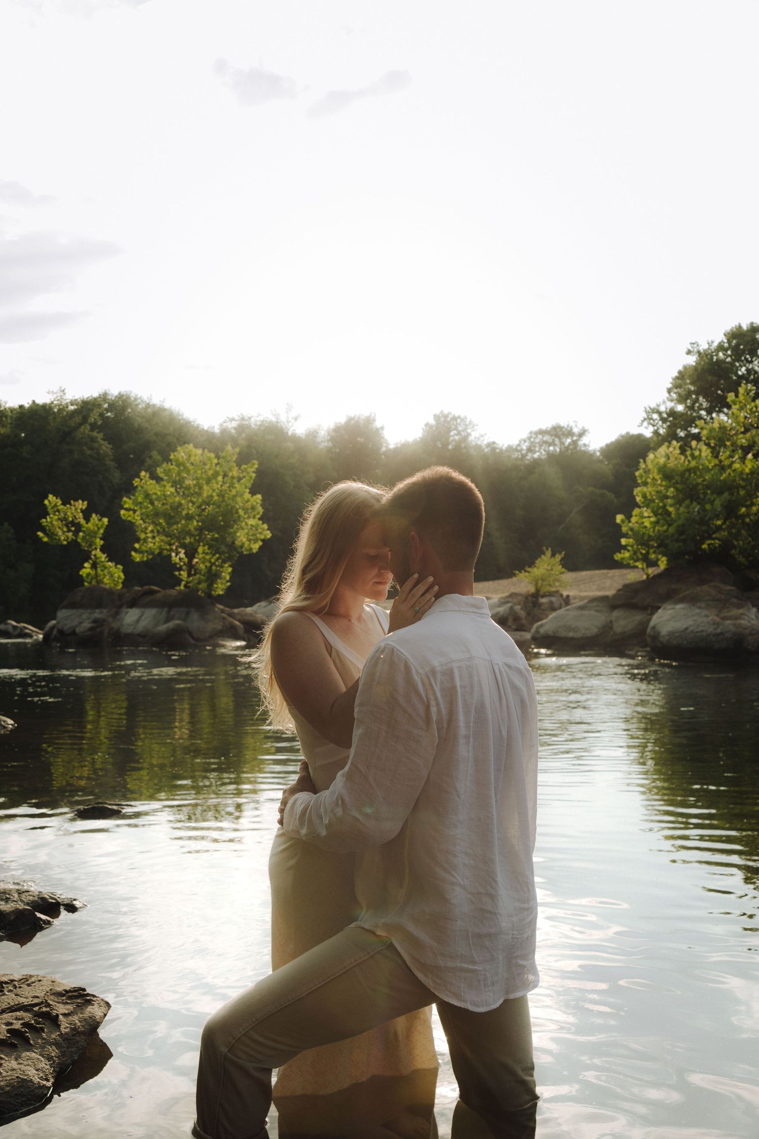 Romantic couple embraces by a serene lake at sunset with golden sunlight streaming through trees in the background.