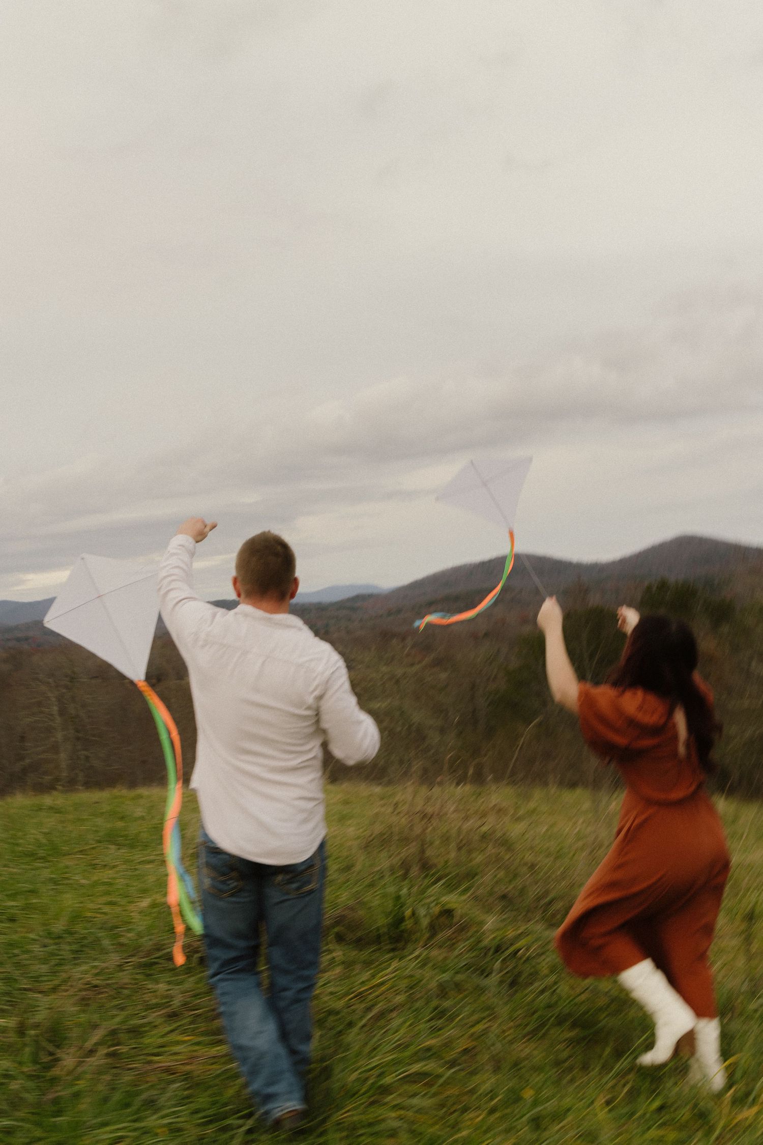 Two people fly colorful kites on a grassy hilltop with mountains in the background on an overcast day.