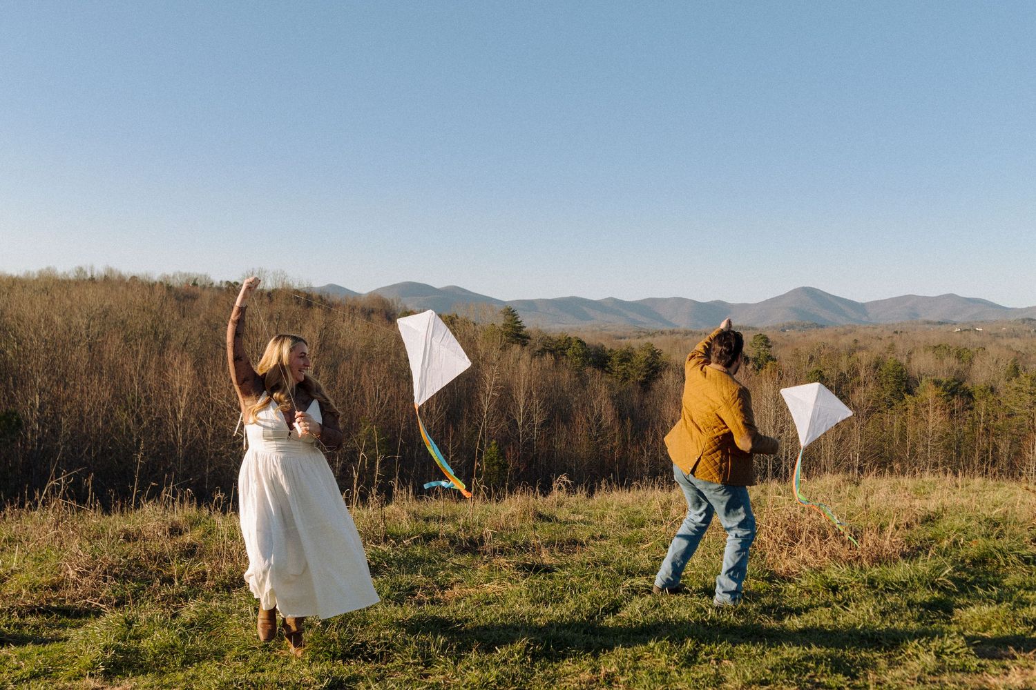 Two people playfully throw blankets in the air while standing on a hilltop overlooking mountains and forest on a sunny day.