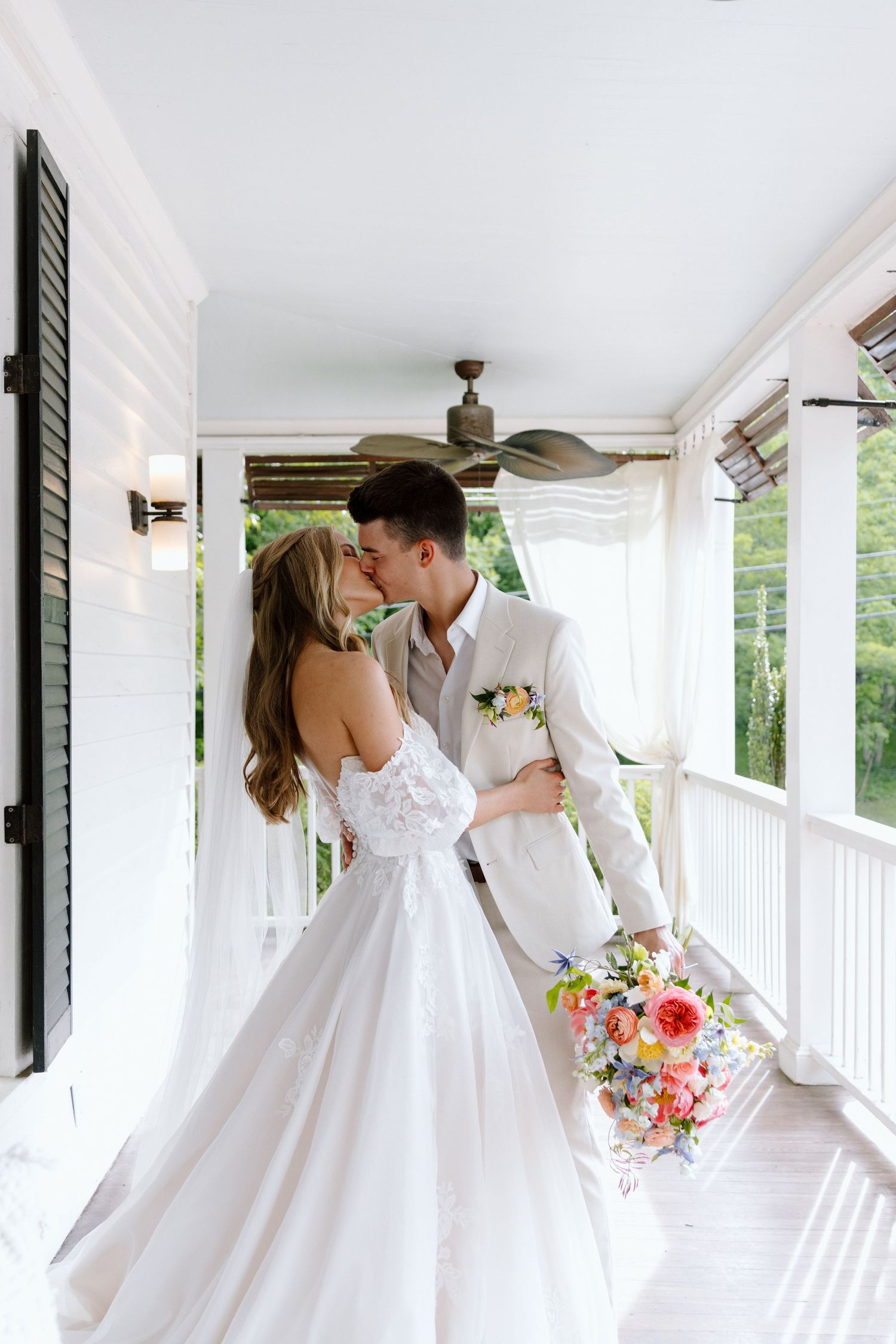Bride and groom share a romantic kiss on a white porch, bride holding a colorful bouquet in a stunning ballgown.