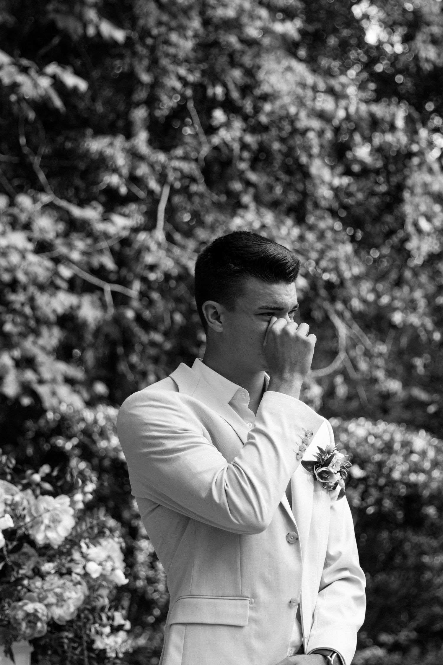Emotional groom in white suit wiping tears at outdoor wedding ceremony with floral backdrop, black and white photo.