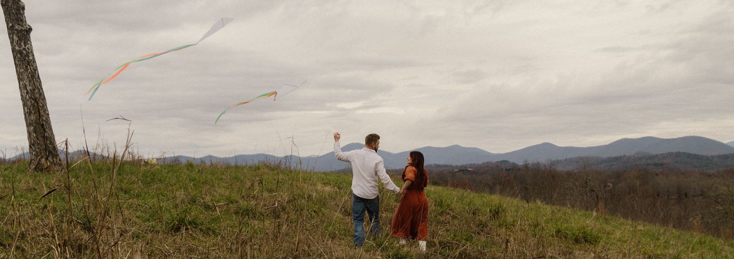 Two people stand on a hill overlooking mountains at sunset, with a dramatic cloudy sky above them.