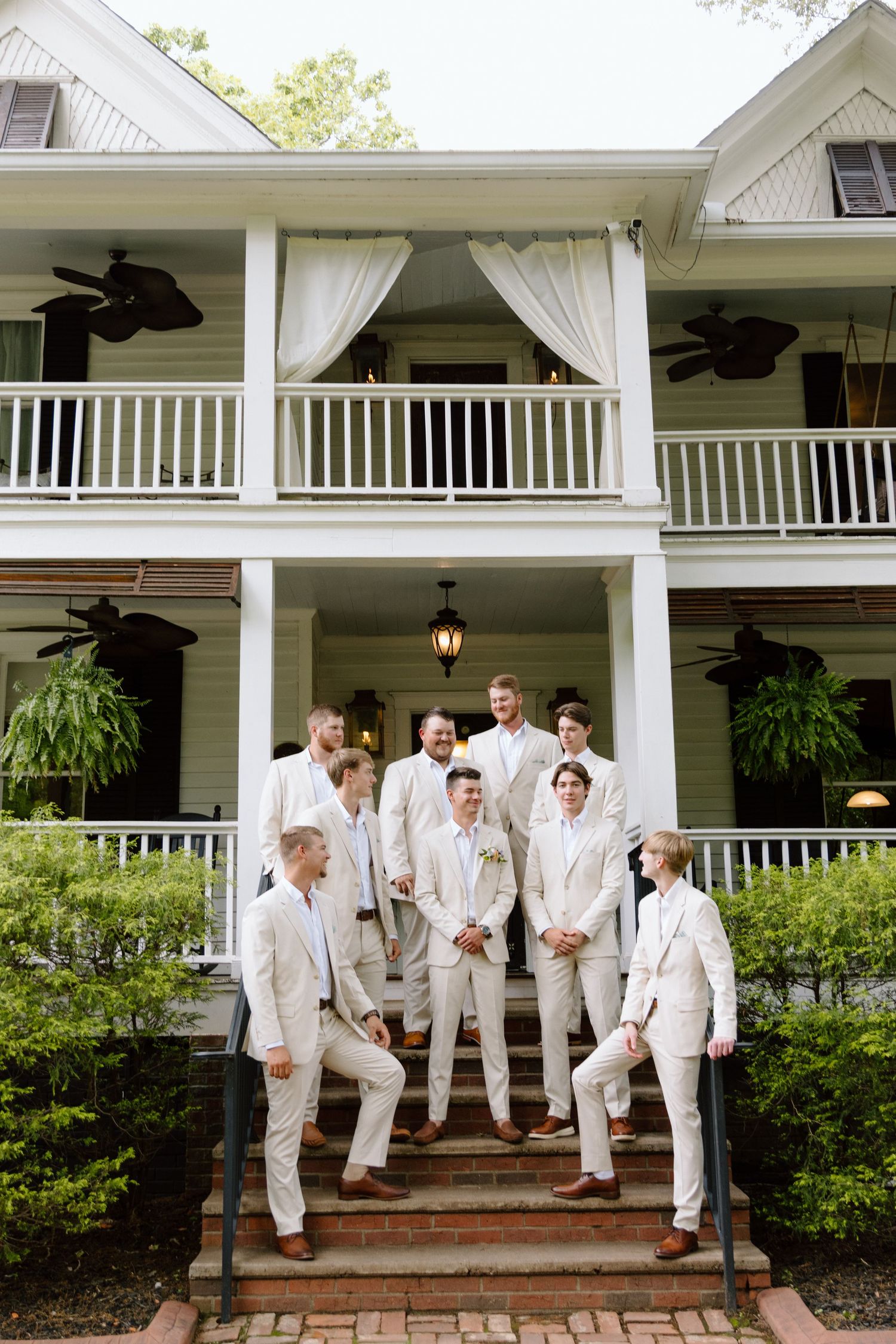 Groomsmen in cream suits pose on brick steps of a charming two-story Southern plantation home with wrap-around porches.