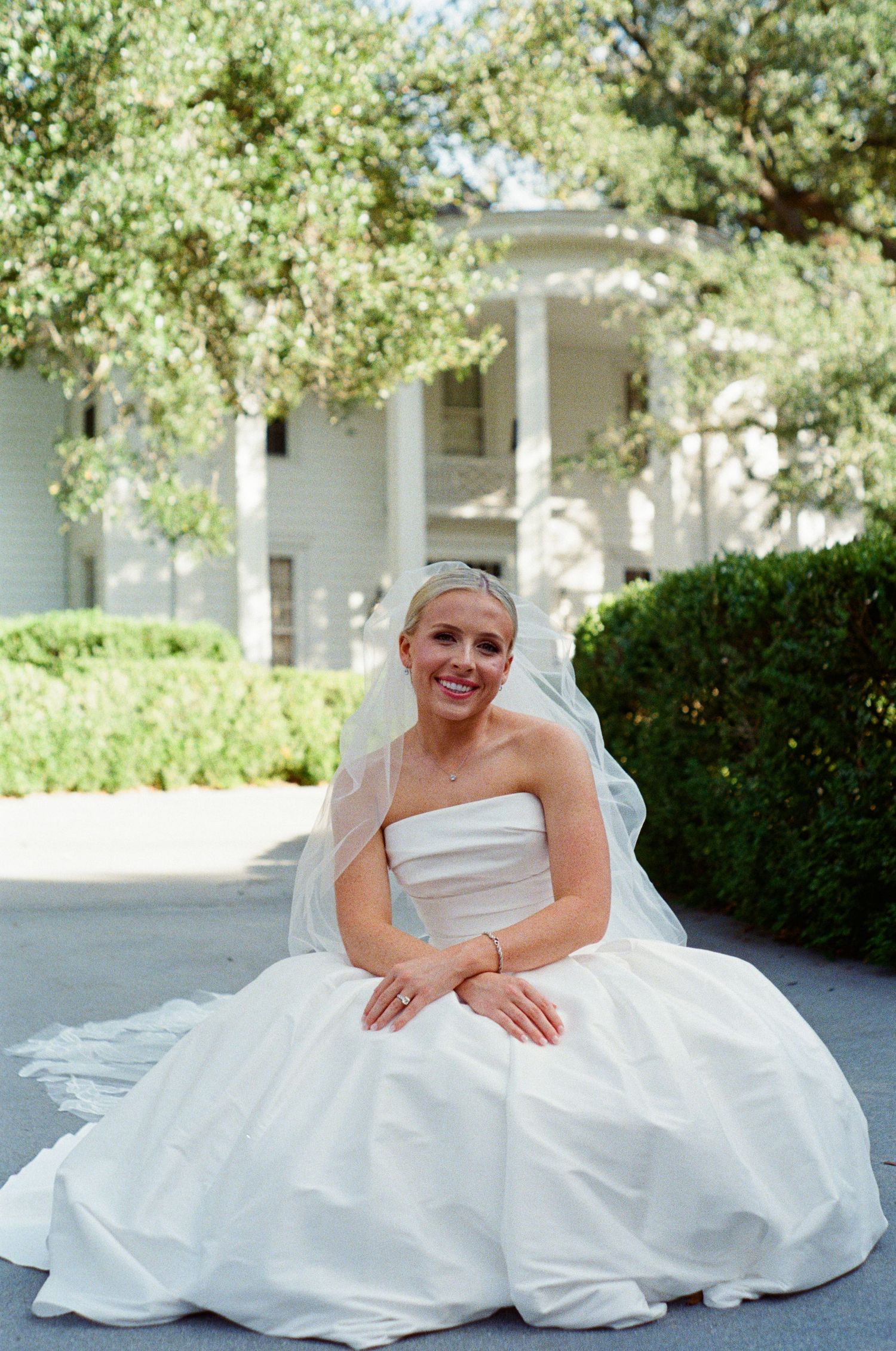 Smiling bride in strapless white ball gown and veil sits outdoors in front of a classic white columned mansion.