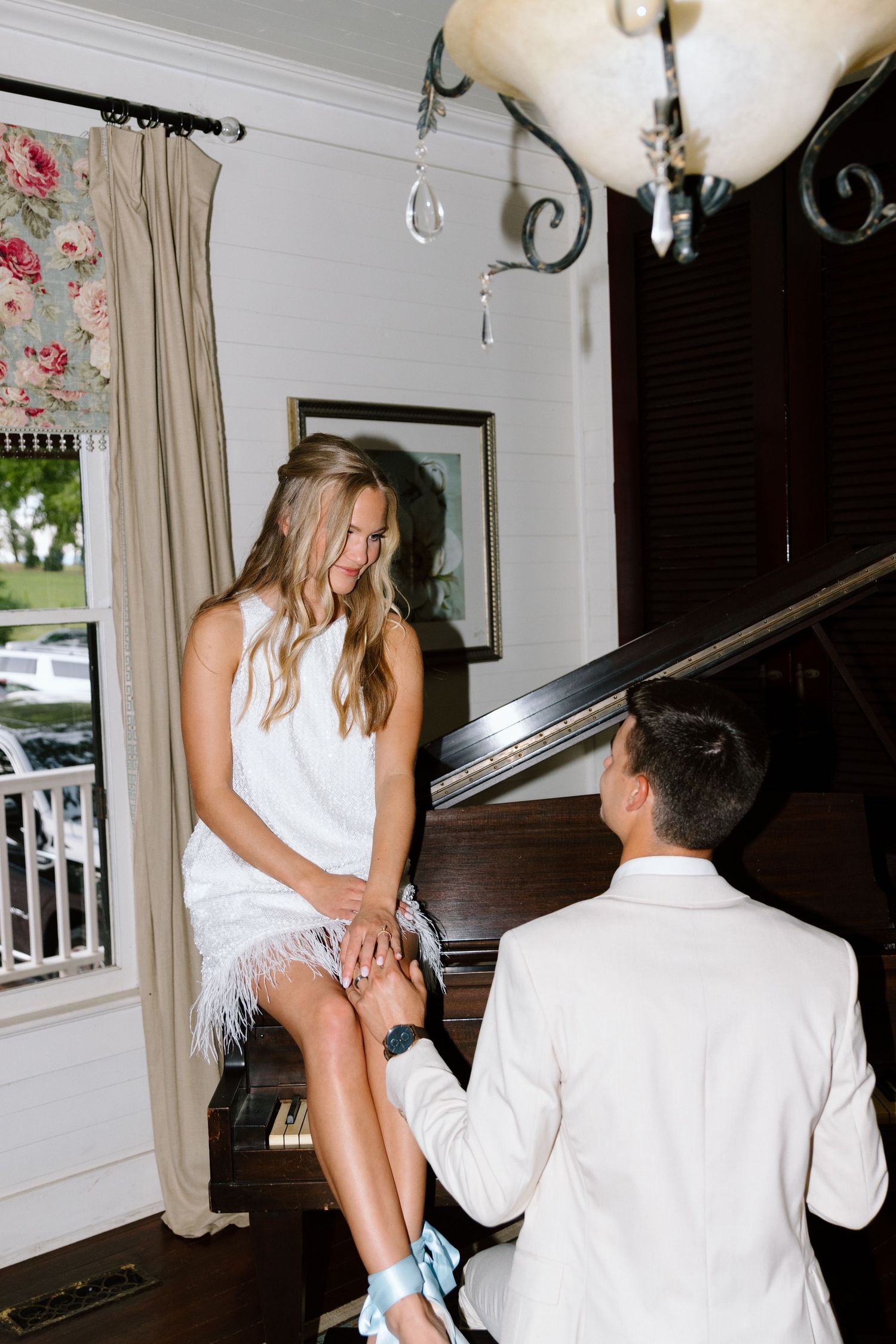 Bride in white feather dress sits on grand piano while groom in white suit plays during wedding reception.
