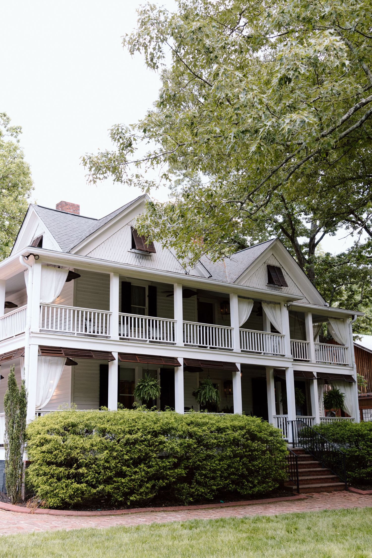 White two-story Southern colonial home with wrap-around porches, black shutters, and lush green trees and shrubs.