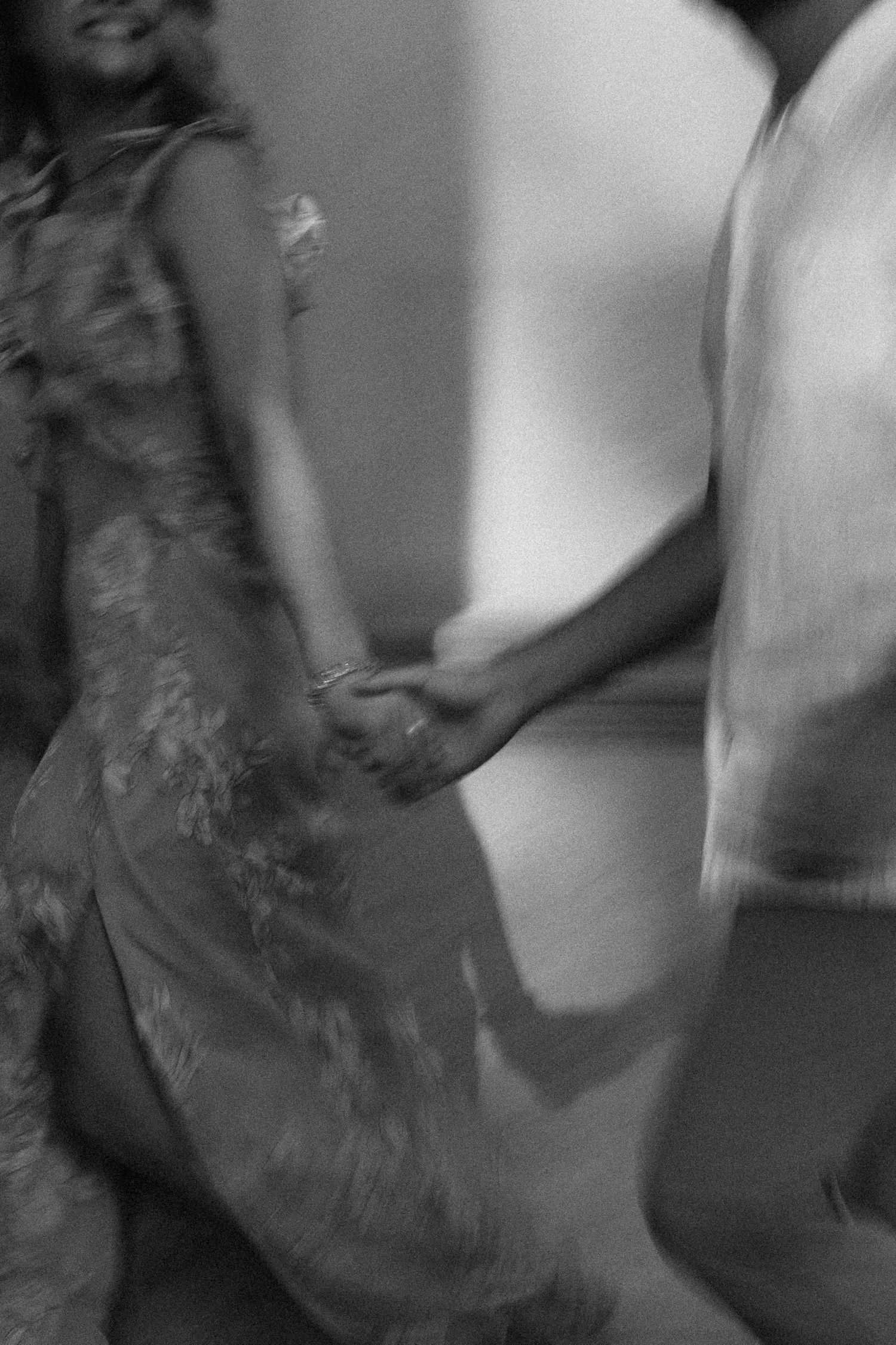 Person dressed in dark clothing stands expressively against a blurred background in dramatic black and white portrait.