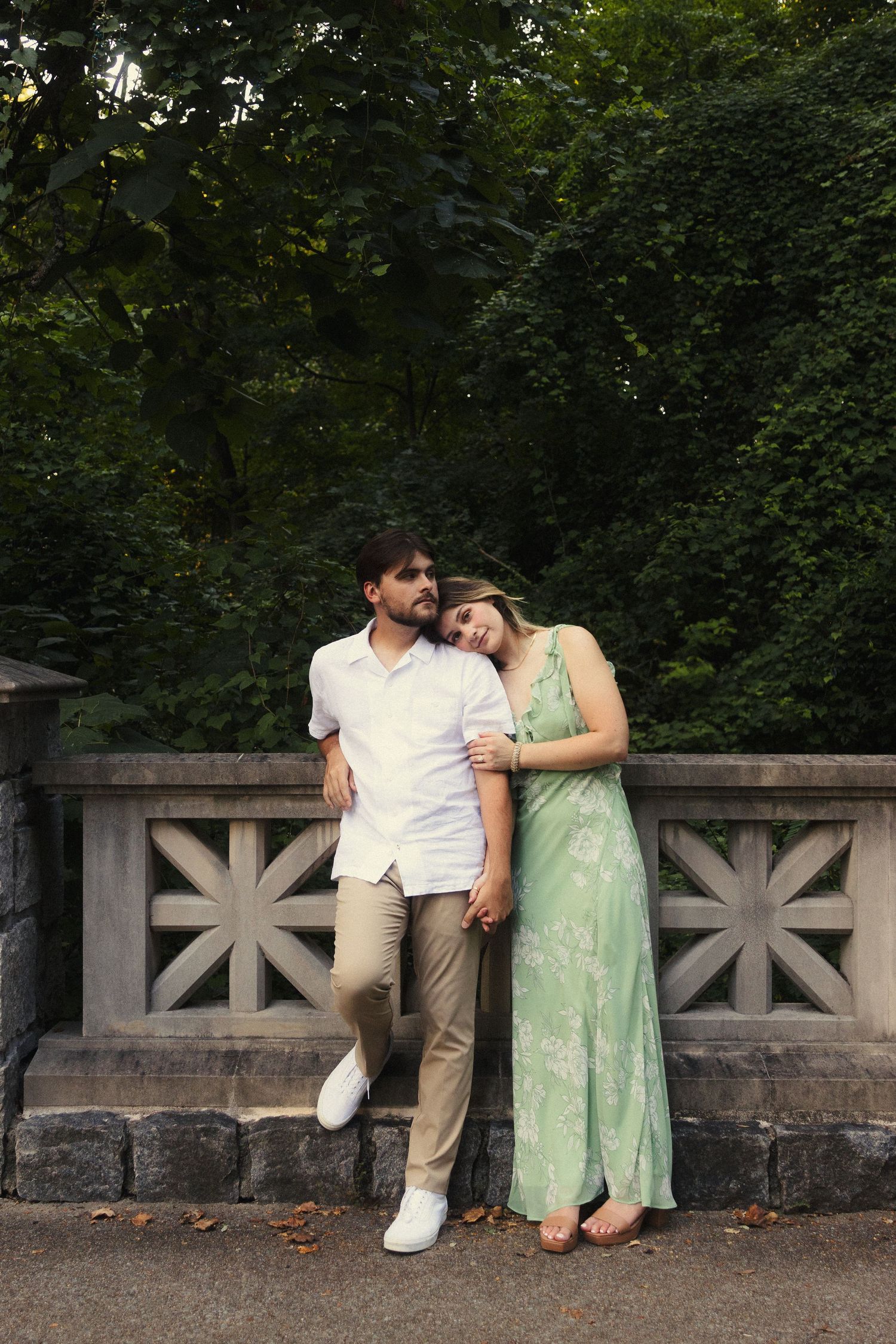 A couple in casual attire share a tender moment while sitting together on a stone wall surrounded by green foliage.