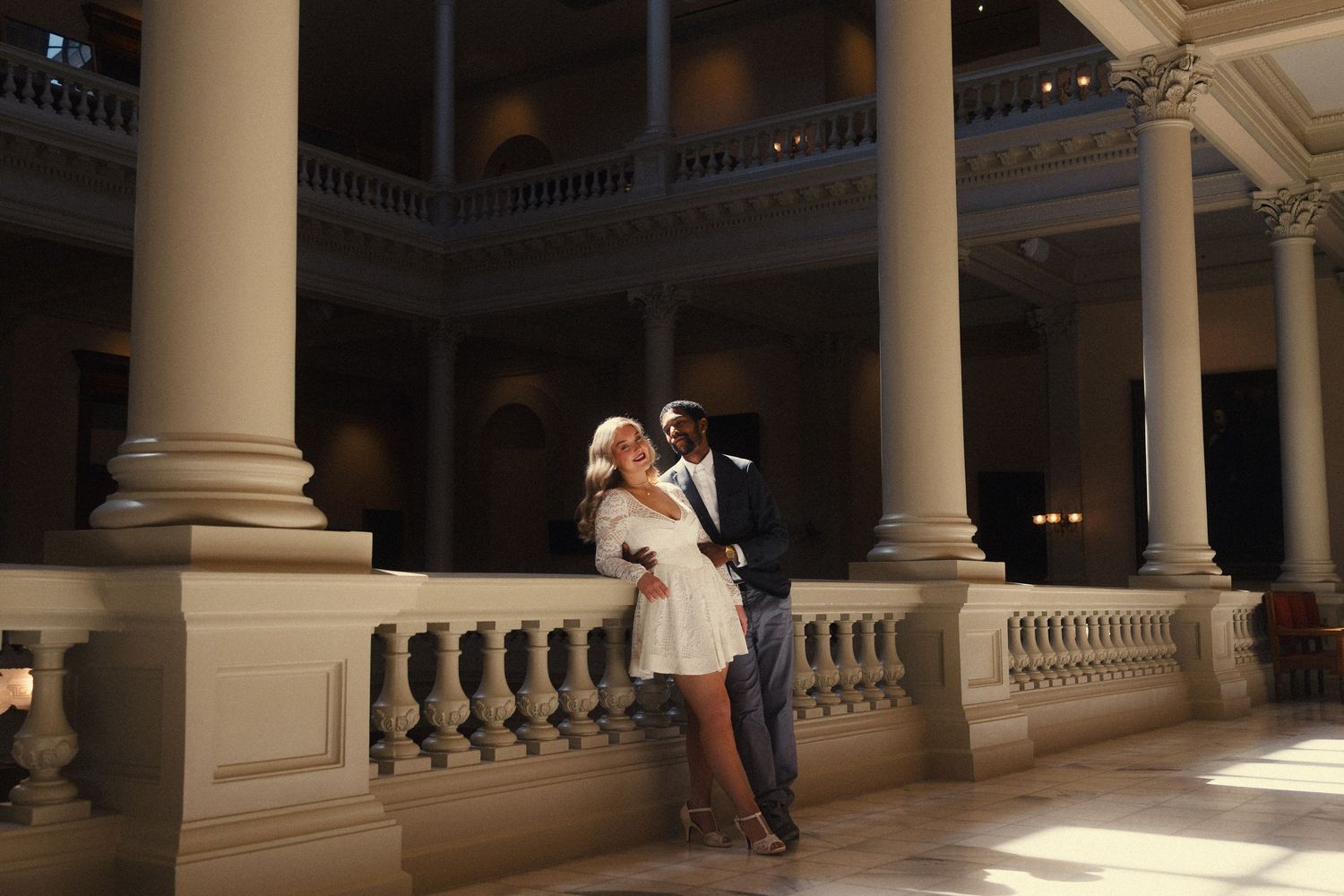 A person leans against a white pillar in an elegant columned gallery with ornate architectural details.