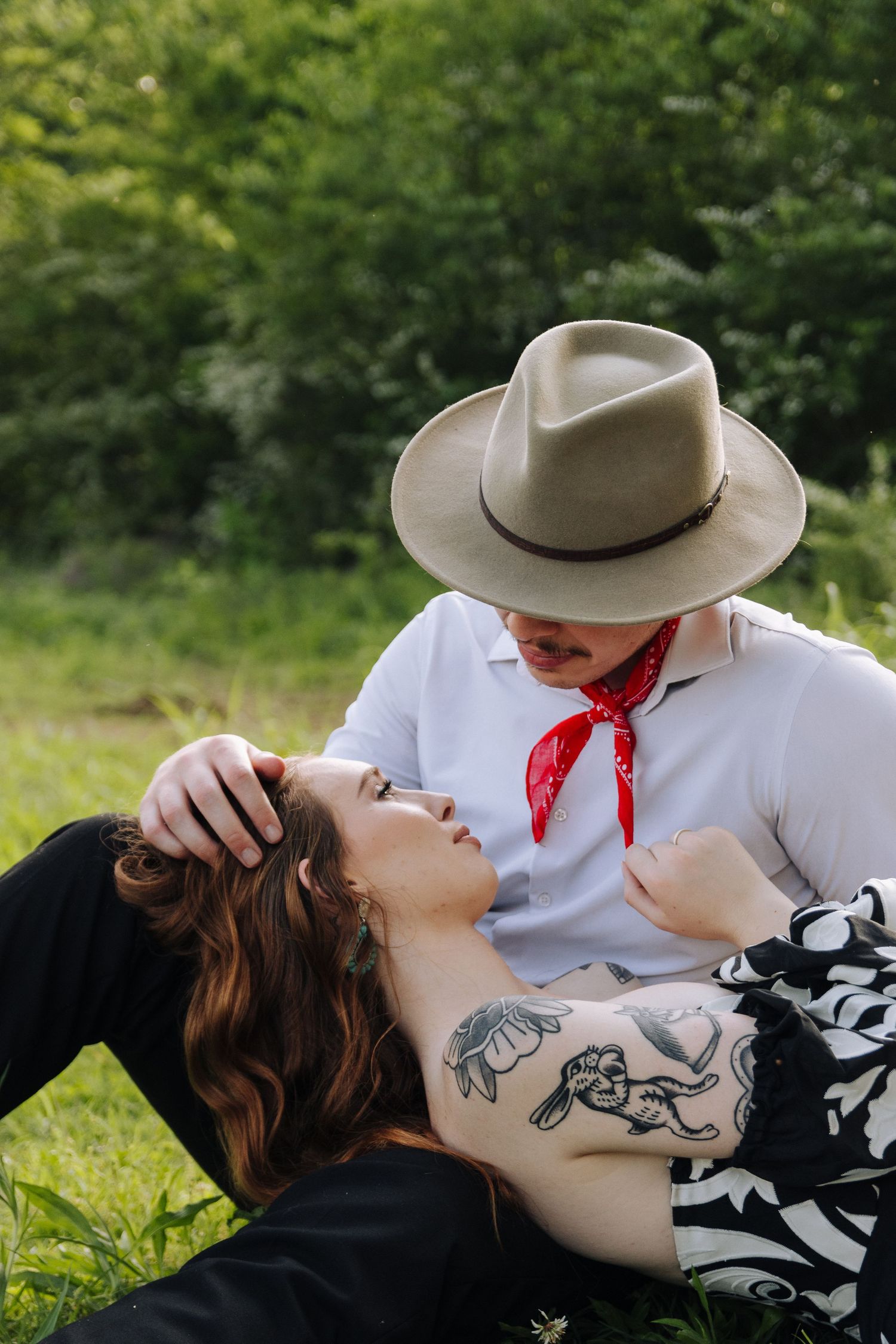 A figure in a cowboy hat and red bandana leans over someone reclining on grass in a romantic photoshoot series.