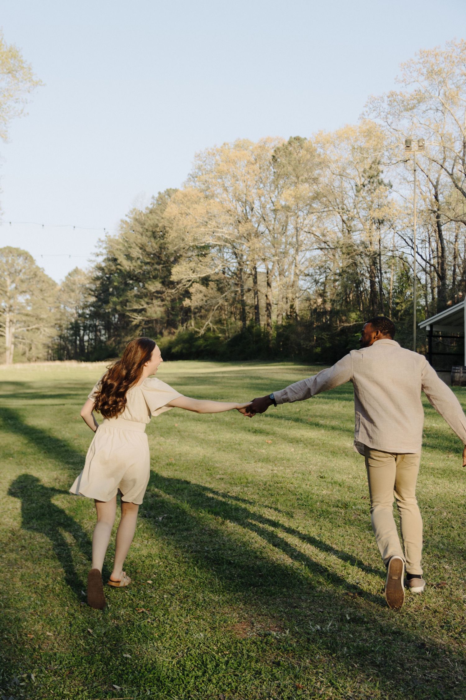A couple in neutral colored outfits holding hands while walking across a sunlit grassy lawn with trees in the background.