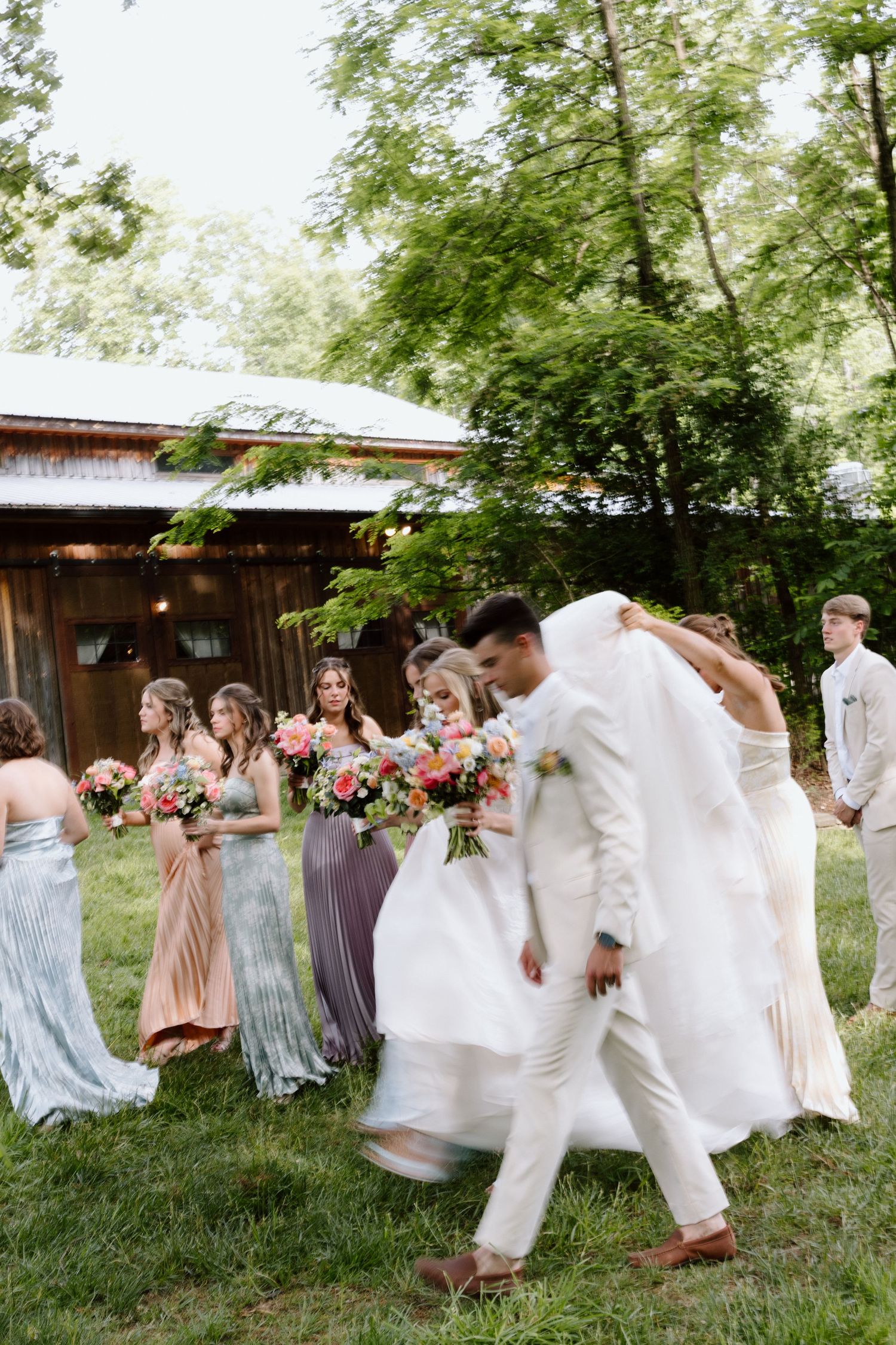 Wedding party walking outdoors, bride in white gown with groom in white suit, bridesmaids in colorful dresses holding floral bouquets.