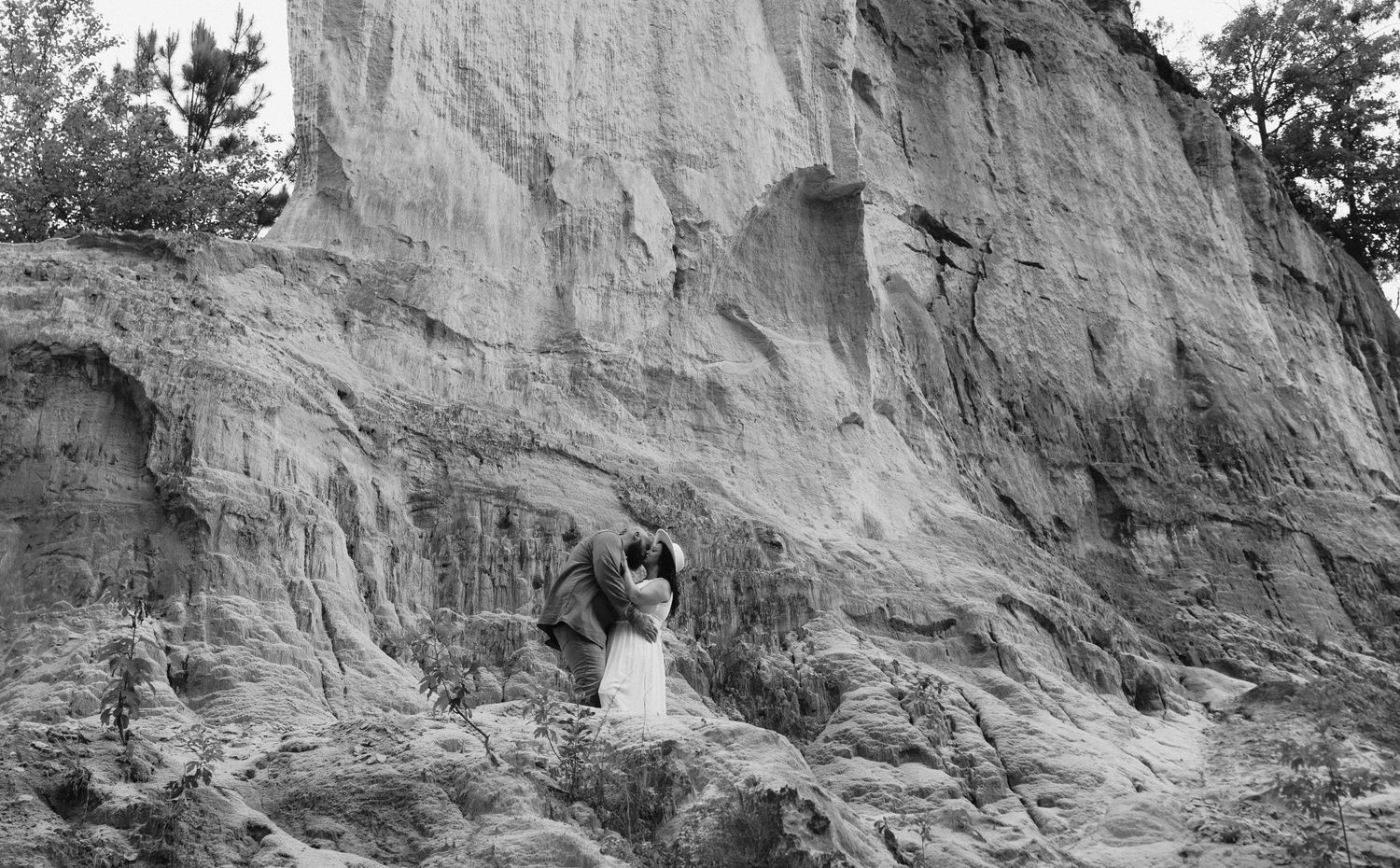 A person stands in front of a towering natural clay or sand cliff face in black and white.