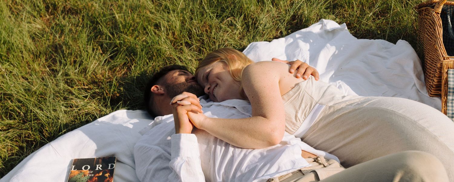 A couple resting on white blankets outdoors in a grassy meadow on a sunny afternoon.