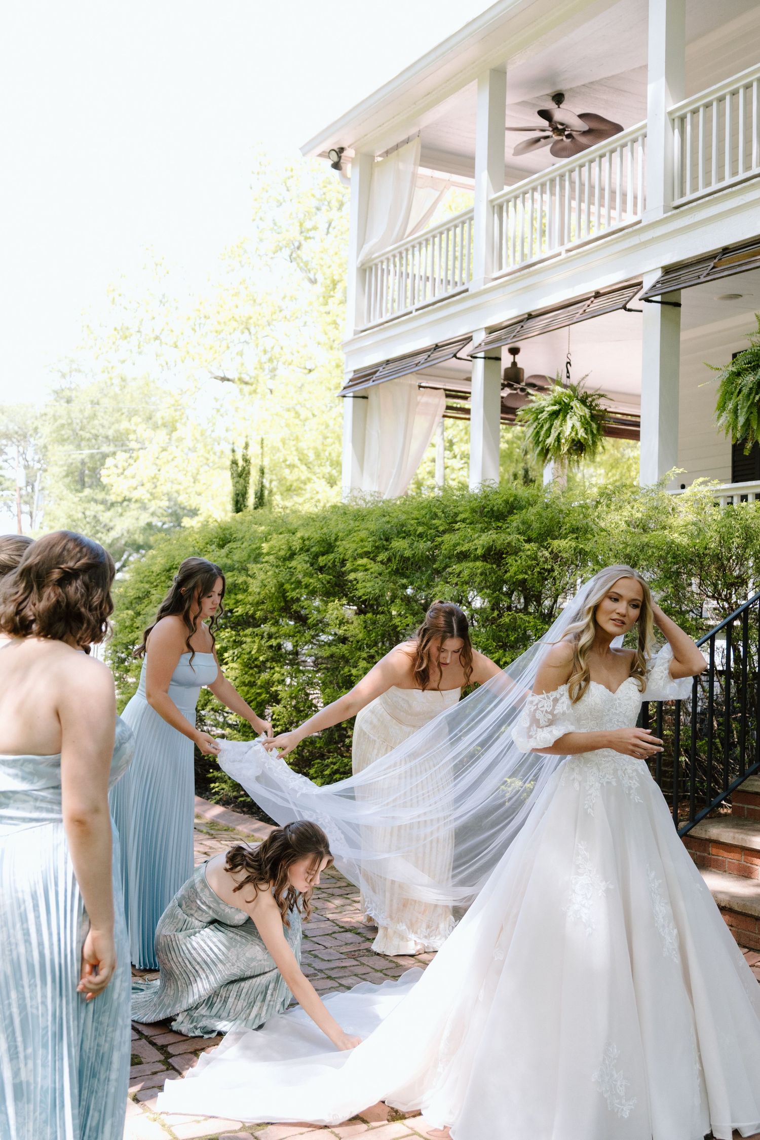 Bridesmaids in blue dresses arrange the veil of a bride in a white gown outside an elegant white mansion.