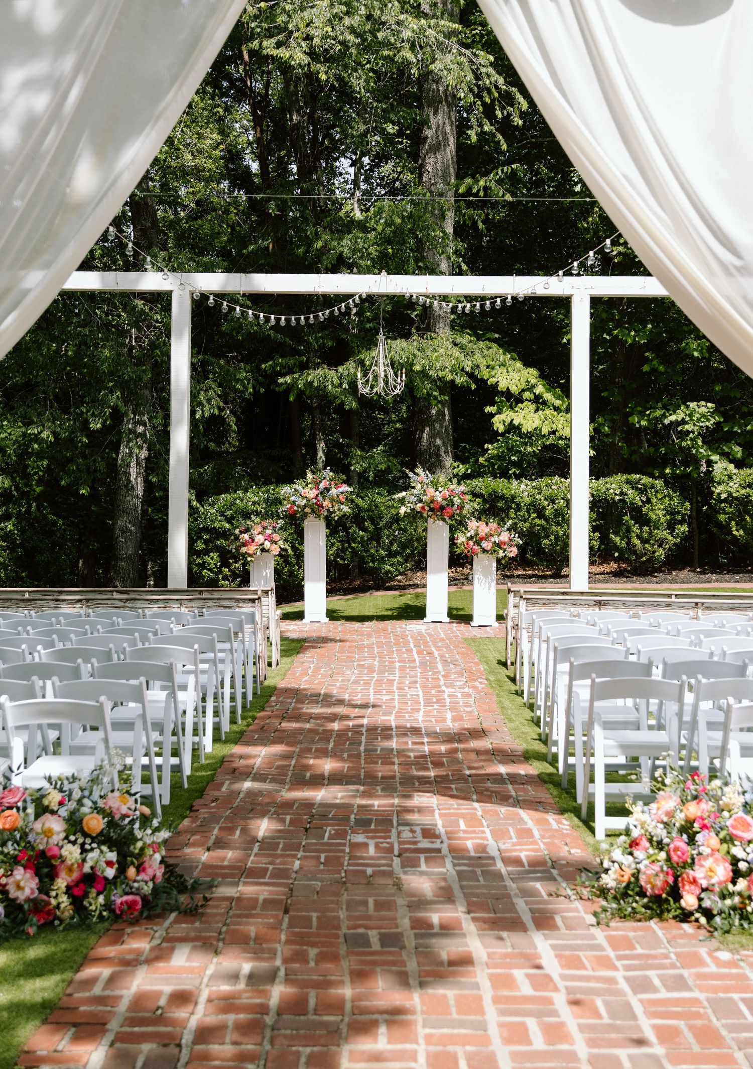Elegant outdoor wedding ceremony with brick aisle, white chairs, floral arrangements, and a white arbor surrounded by lush greenery.