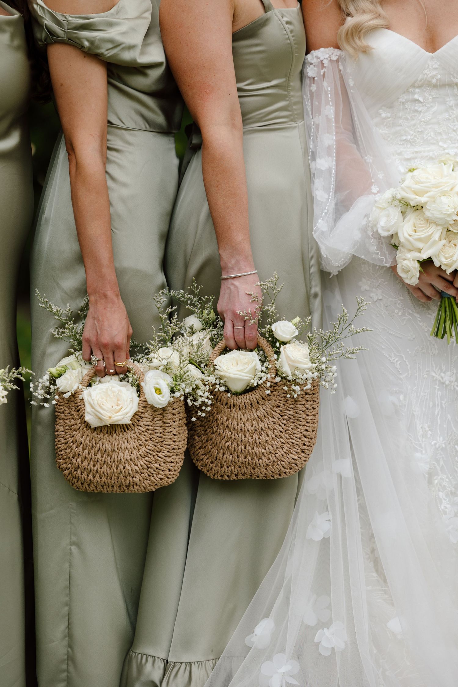 Bridesmaids in sage green dresses hold woven straw bags filled with white roses and baby's breath flowers.