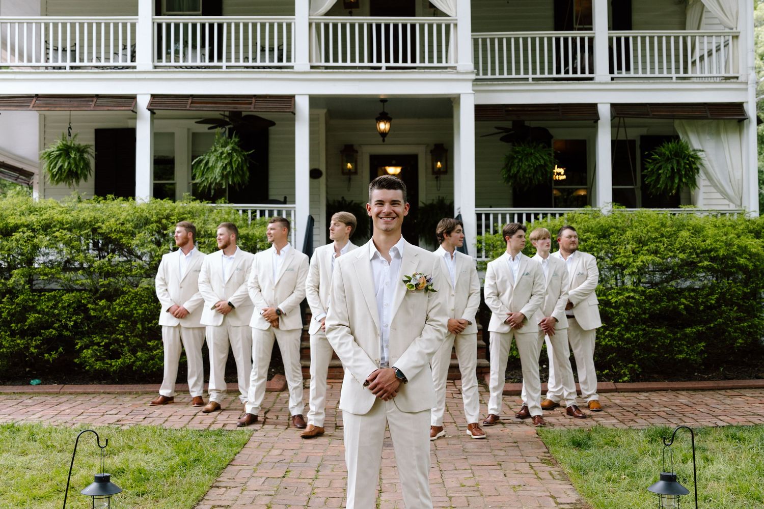 Groom in white linen suit stands before groomsmen in matching attire outside an elegant Southern estate.