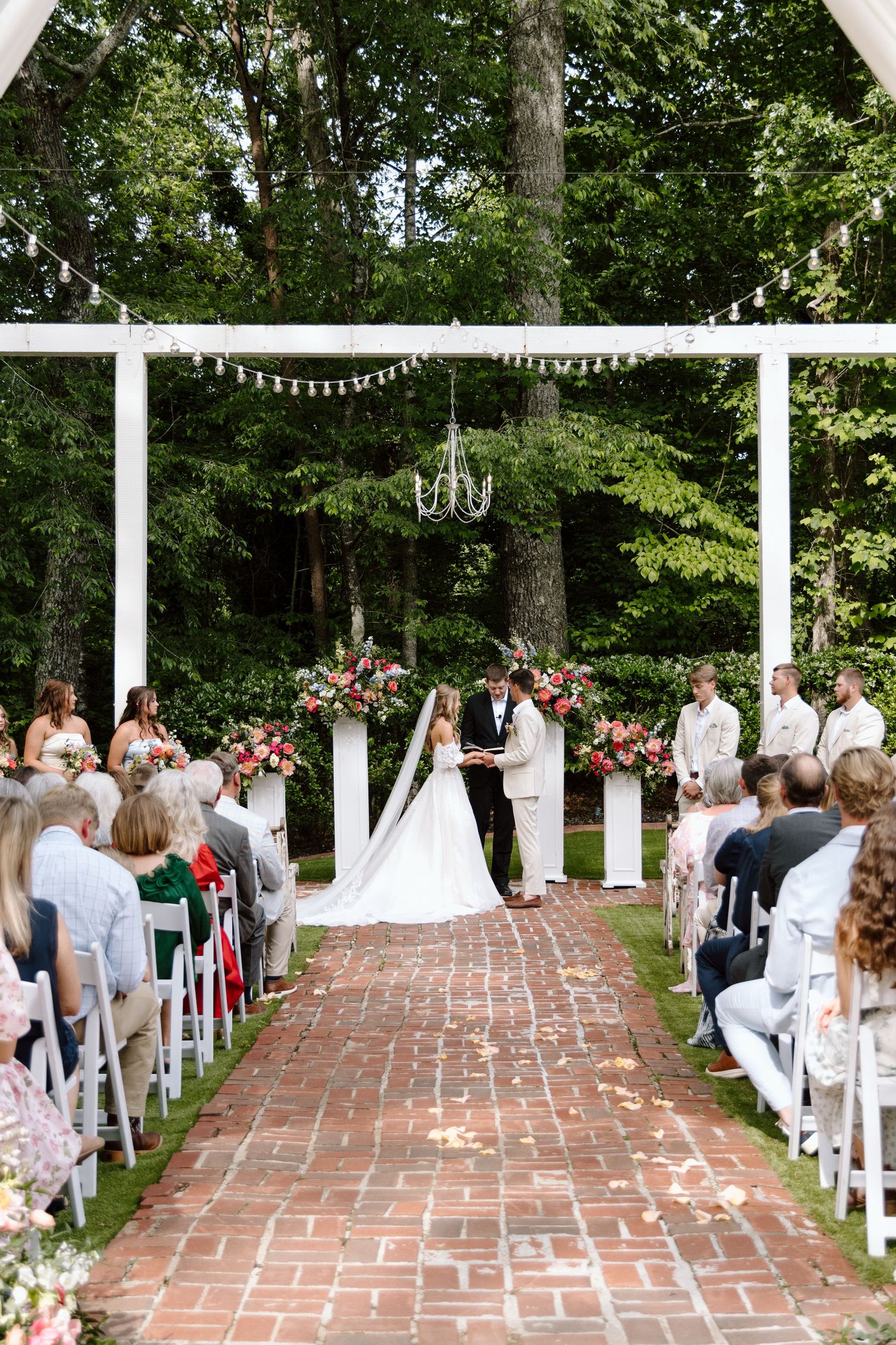 Outdoor wedding ceremony on brick aisle under white pergola with chandelier, floral arrangements, and lush green trees backdrop.