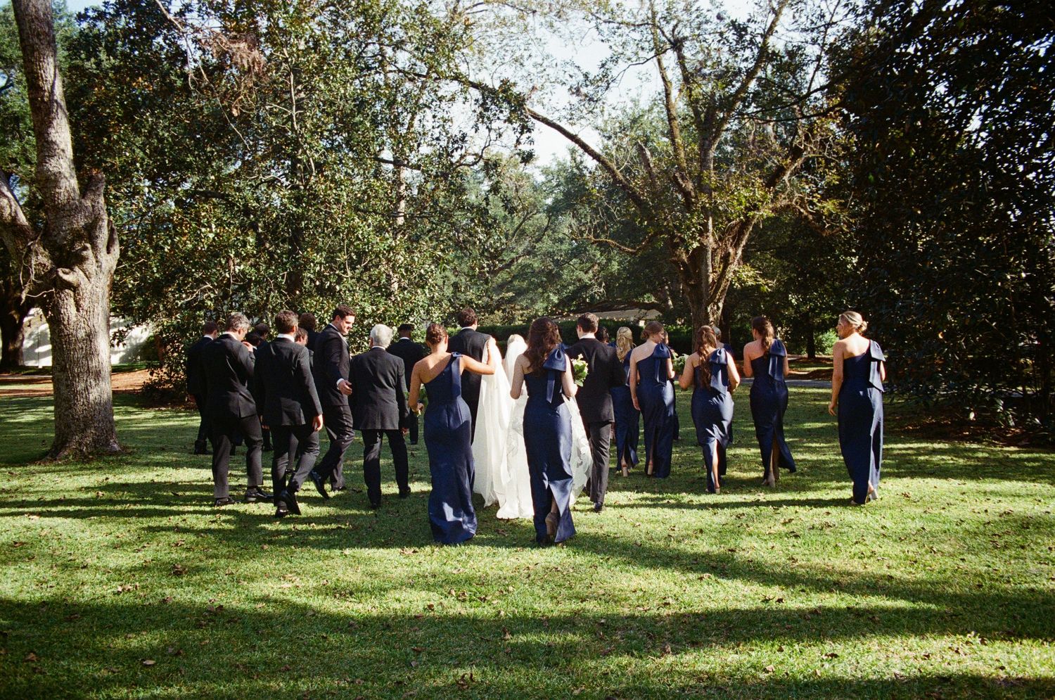 Wedding party in navy attire gathered on lush green lawn surrounded by trees during outdoor ceremony.