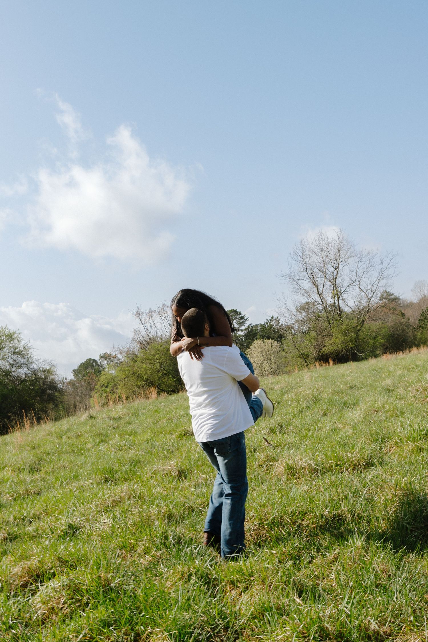 Person in white sweater and jeans standing on grassy hillside under blue sky with white clouds.