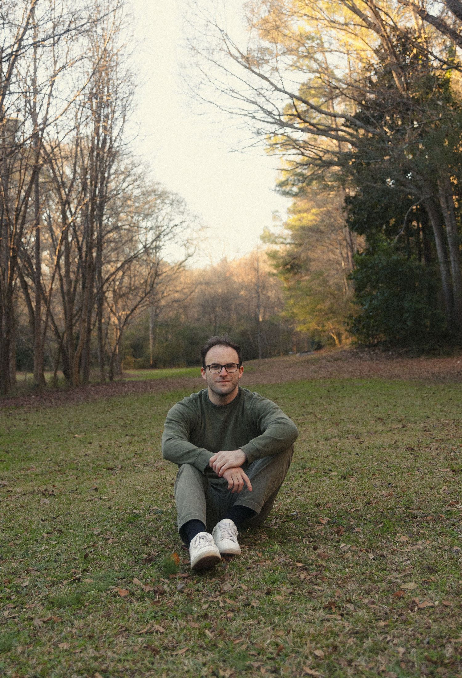 A person in a green sweater sits on grass in a park during autumn as the sun sets behind bare trees.