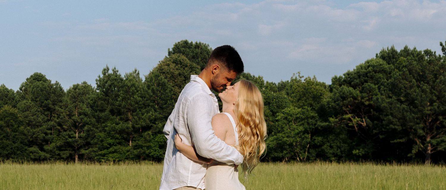 A couple shares a romantic kiss in a scenic outdoor field during sunset.