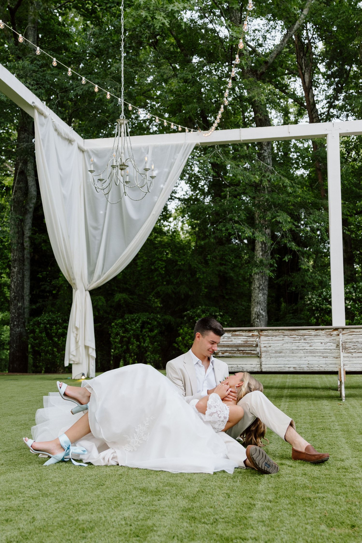 Bride and groom sitting together on green grass beneath a white draped wedding arch with chandelier outdoors.