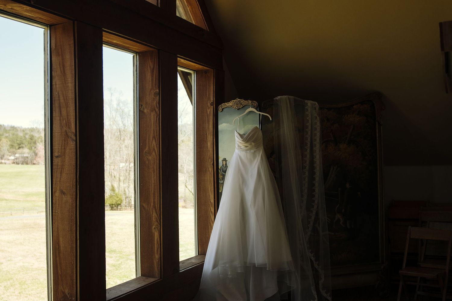 Elegant white wedding dress hanging near large wooden-framed windows overlooking a green field.
