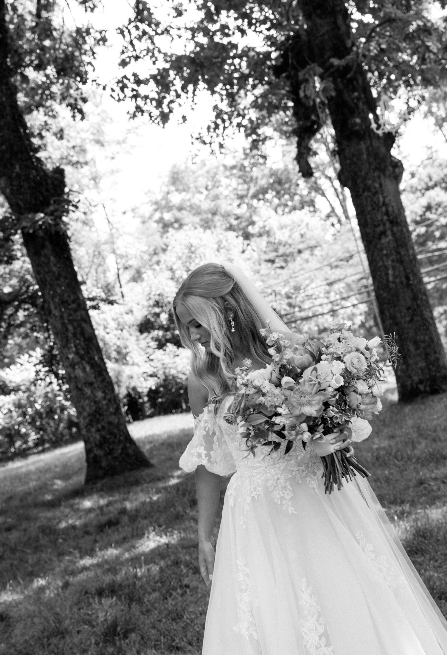 Black and white photo of a bride in a lace gown holding a floral bouquet outdoors among tall trees.