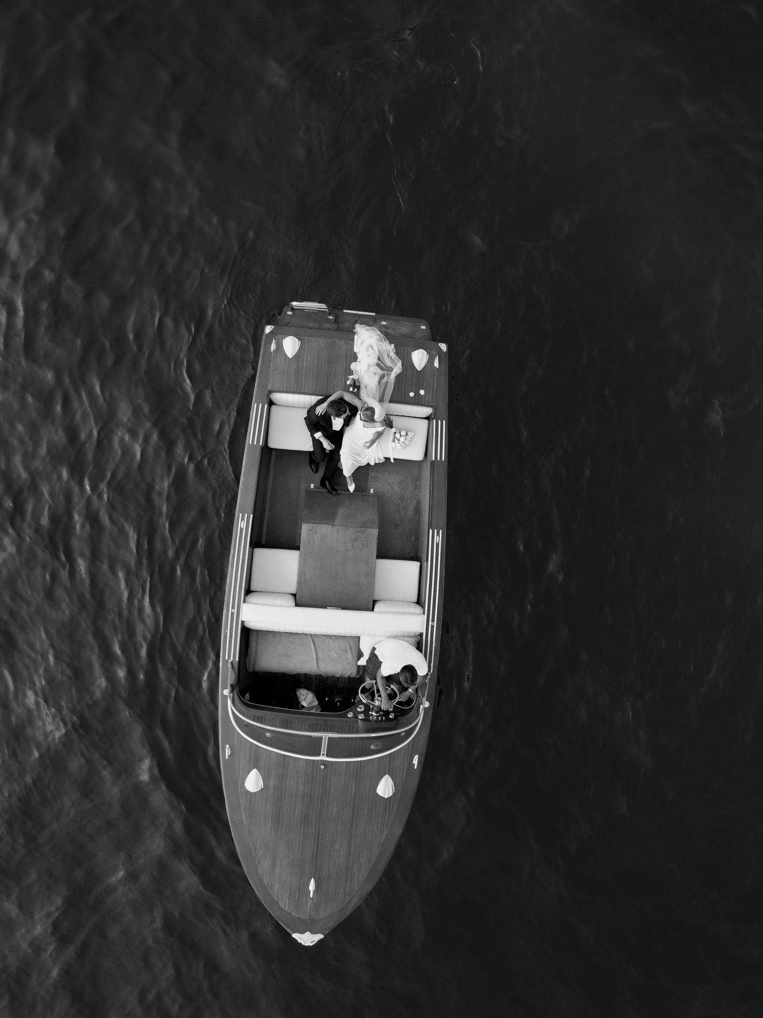 Aerial black and white photo of a wooden speedboat on dark water with two people standing on deck.