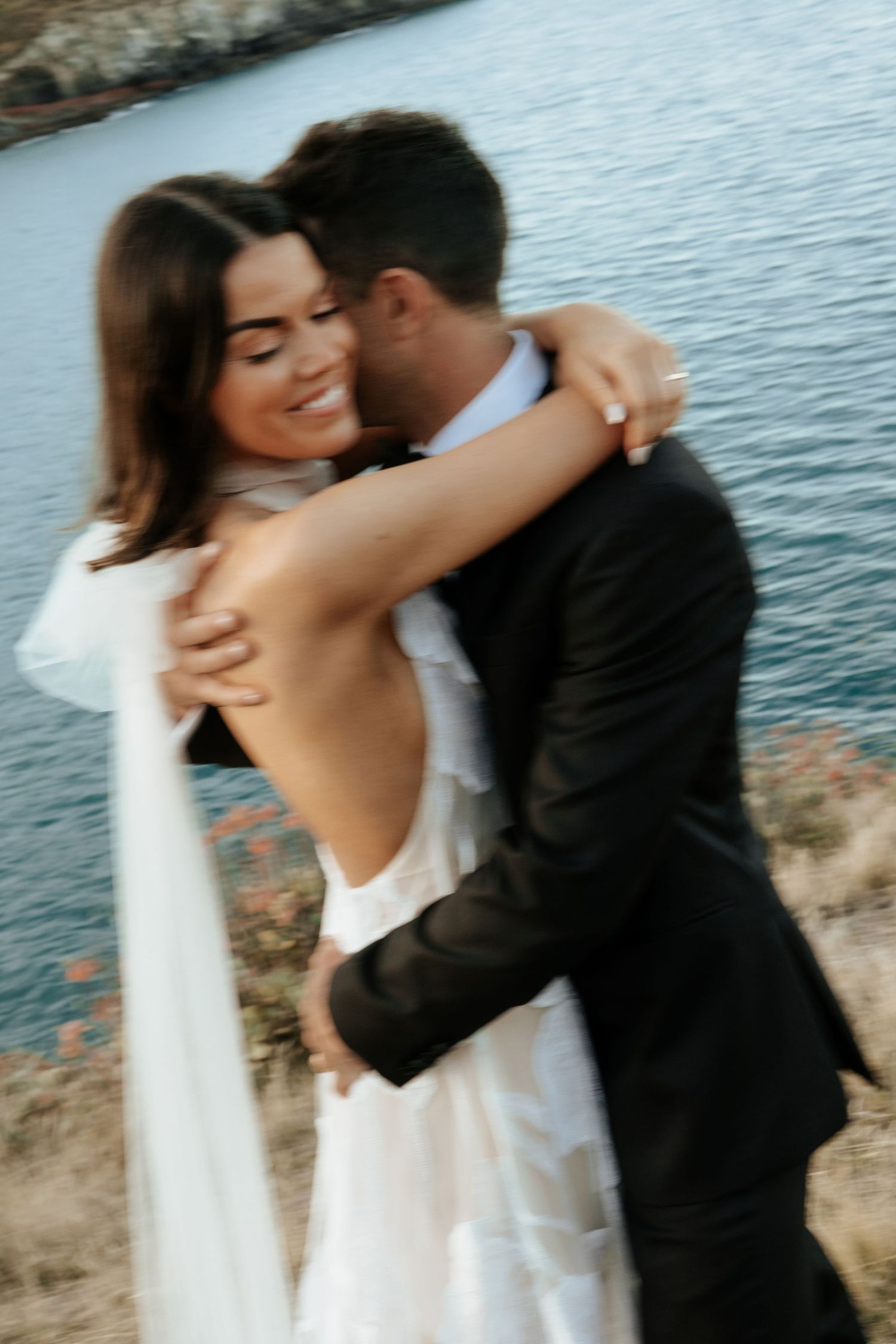 A bride in a backless white gown embraces a groom in a black suit by the ocean.