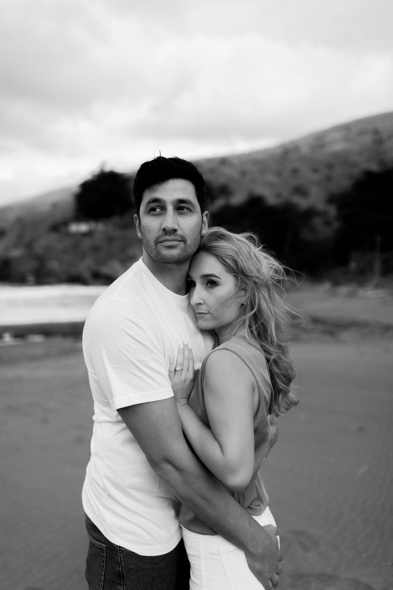 Black and white photo of a couple embracing on a beach, woman hugging man from behind with hills in background.