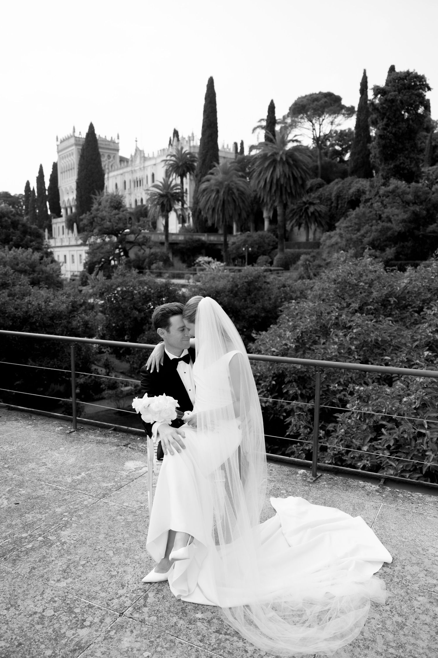 Black and white photo of bride and groom embracing on terrace with elegant villa and lush gardens in background.
