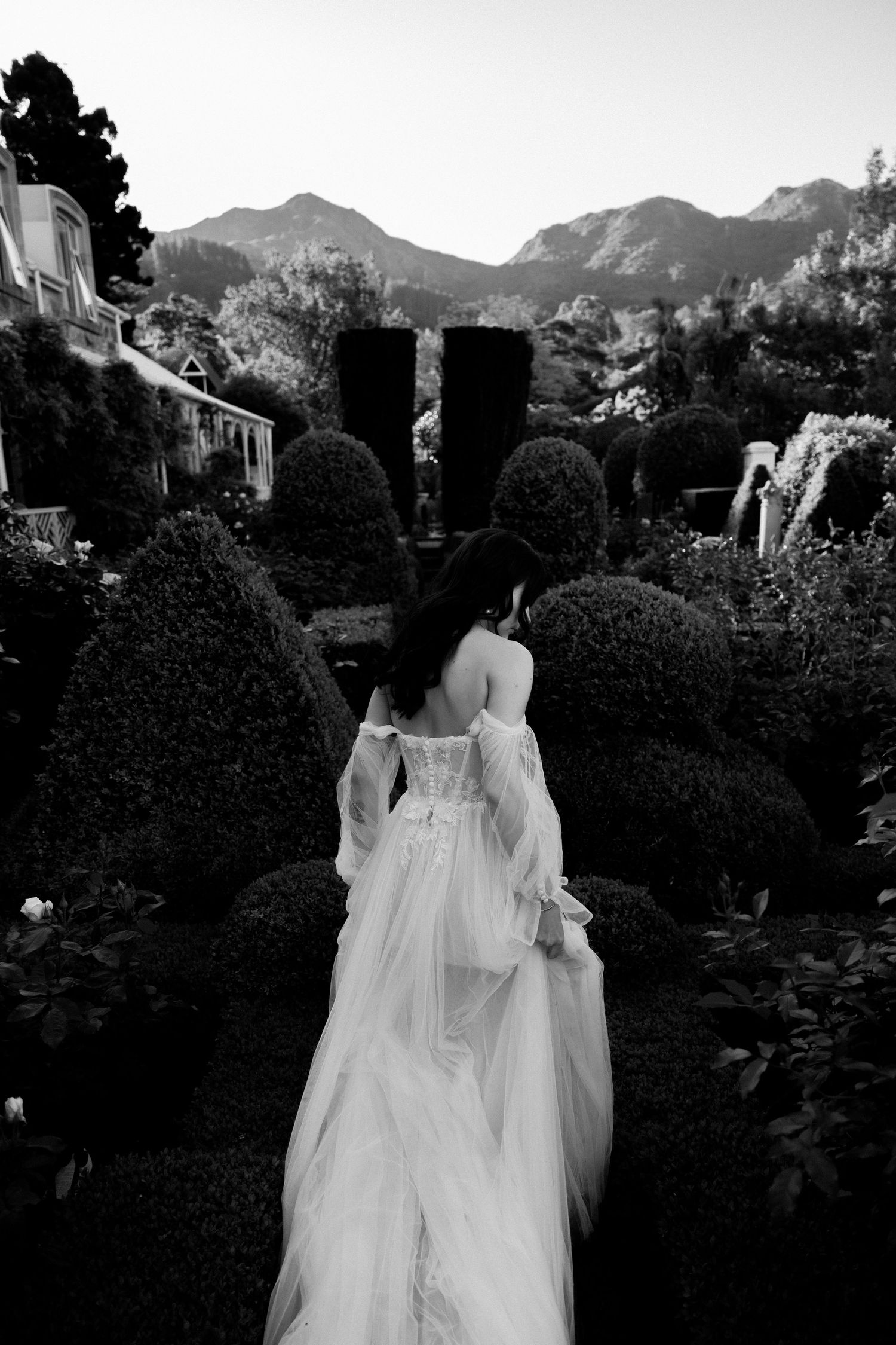 A flowing white wedding dress in a formal garden with manicured topiaries and mountains in the background.