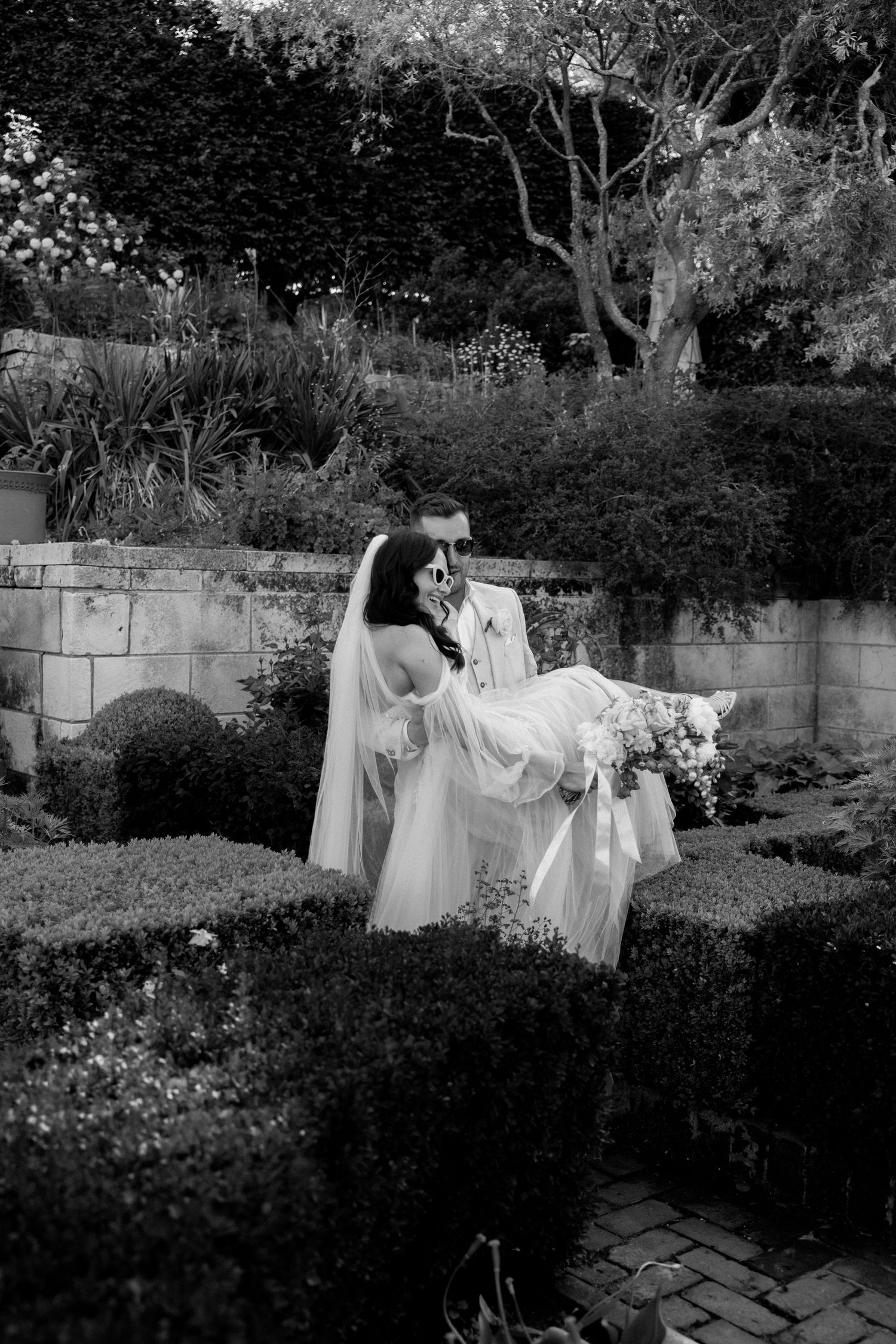 A romantic black and white garden wedding photo with a couple in elegant attire amongst stone walls and lush foliage.