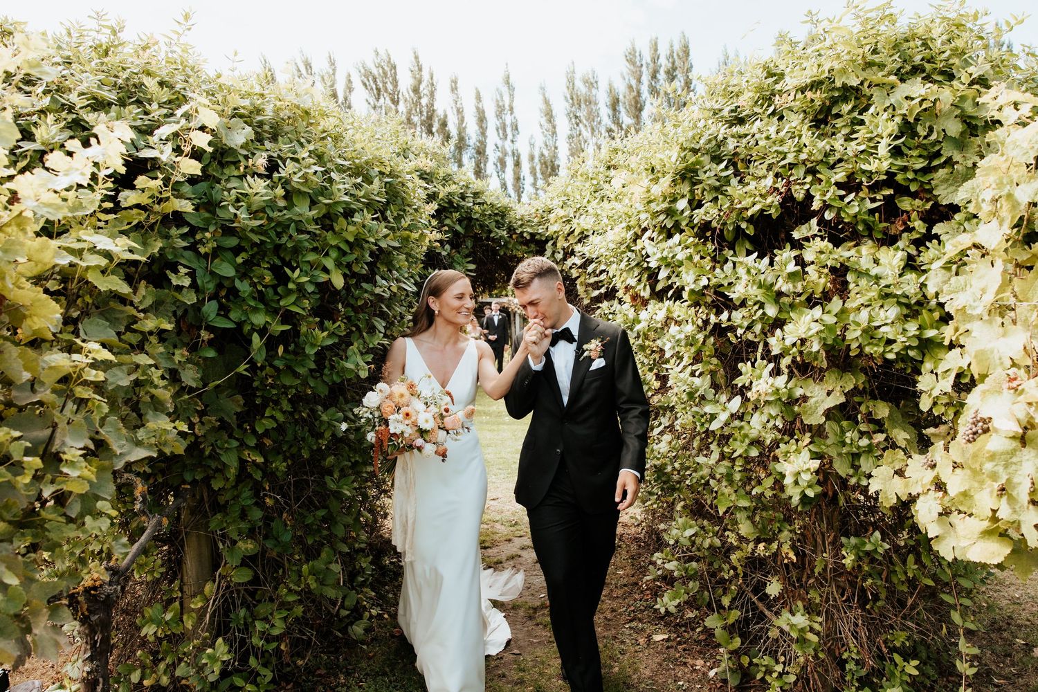 A couple walks together through a lush garden pathway lined with flowering bushes and trees on a sunny day.
