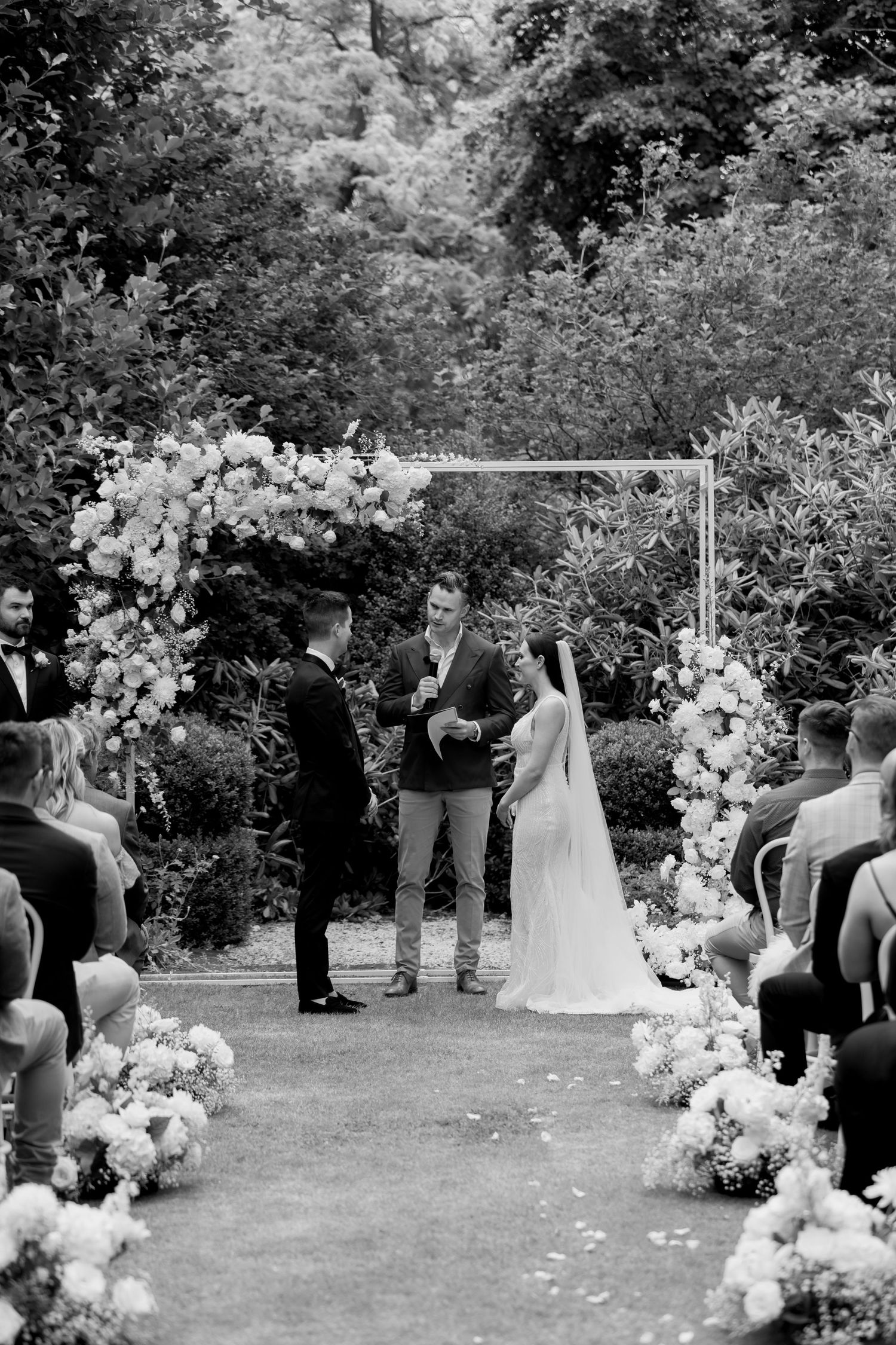 Black and white photo of an outdoor garden wedding ceremony with floral arch decorations.