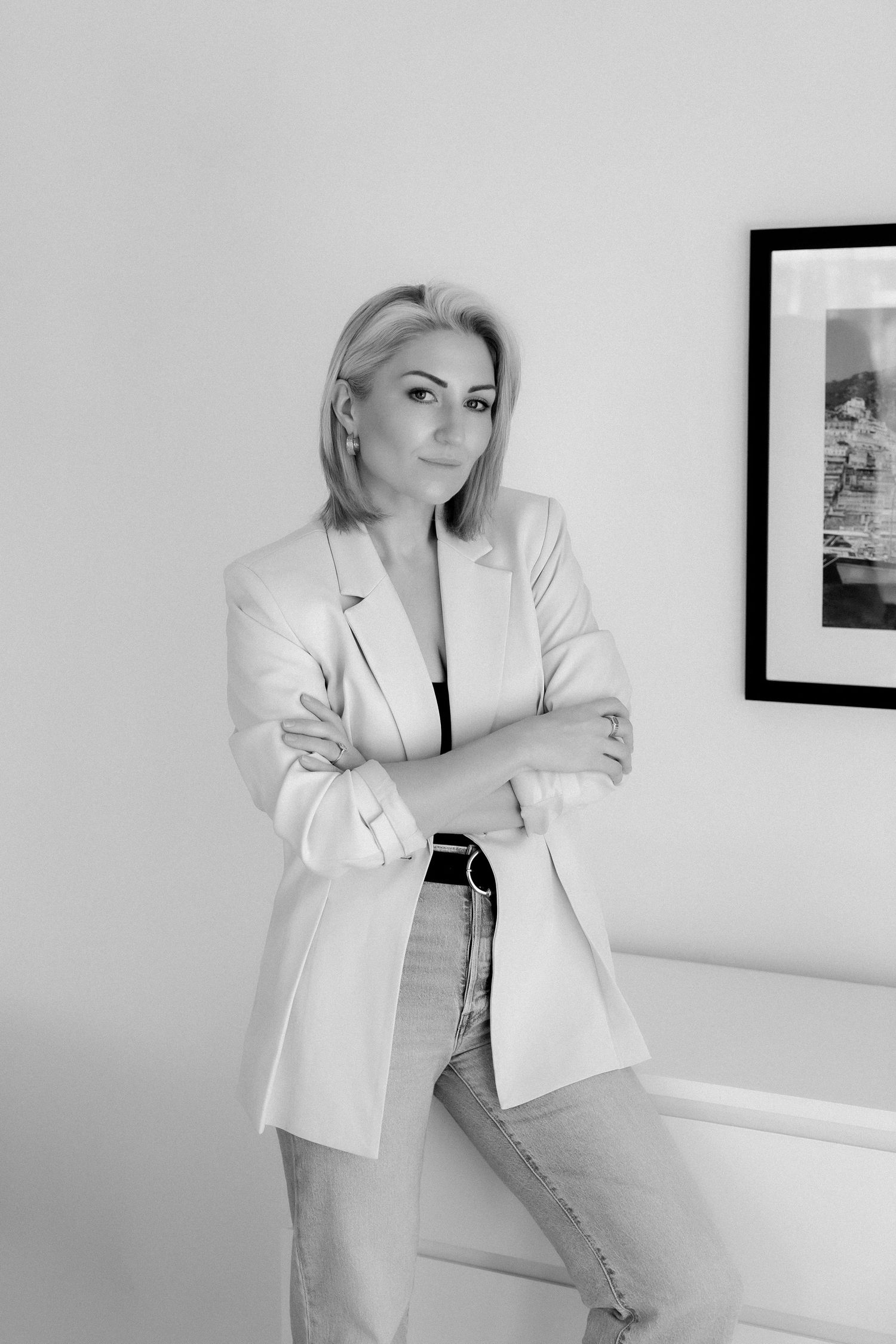 Black and white photo of a confident blonde woman in a blazer and jeans, arms crossed, leaning against a wall.