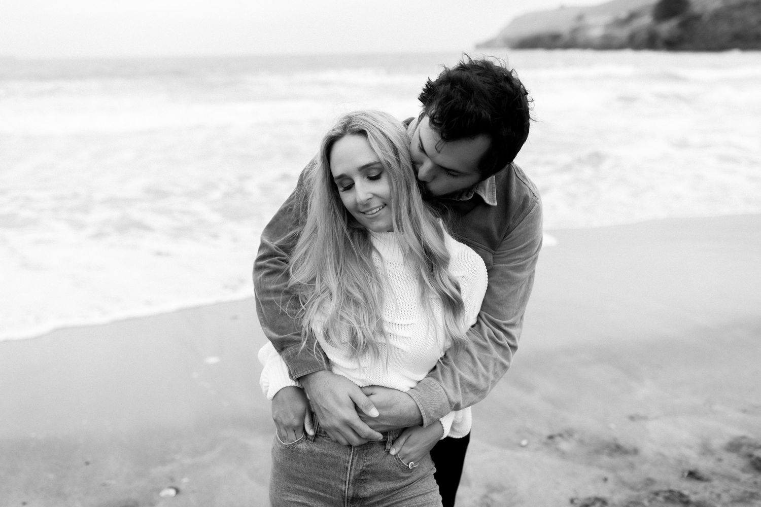 A couple shares an intimate moment on a beach in black and white.